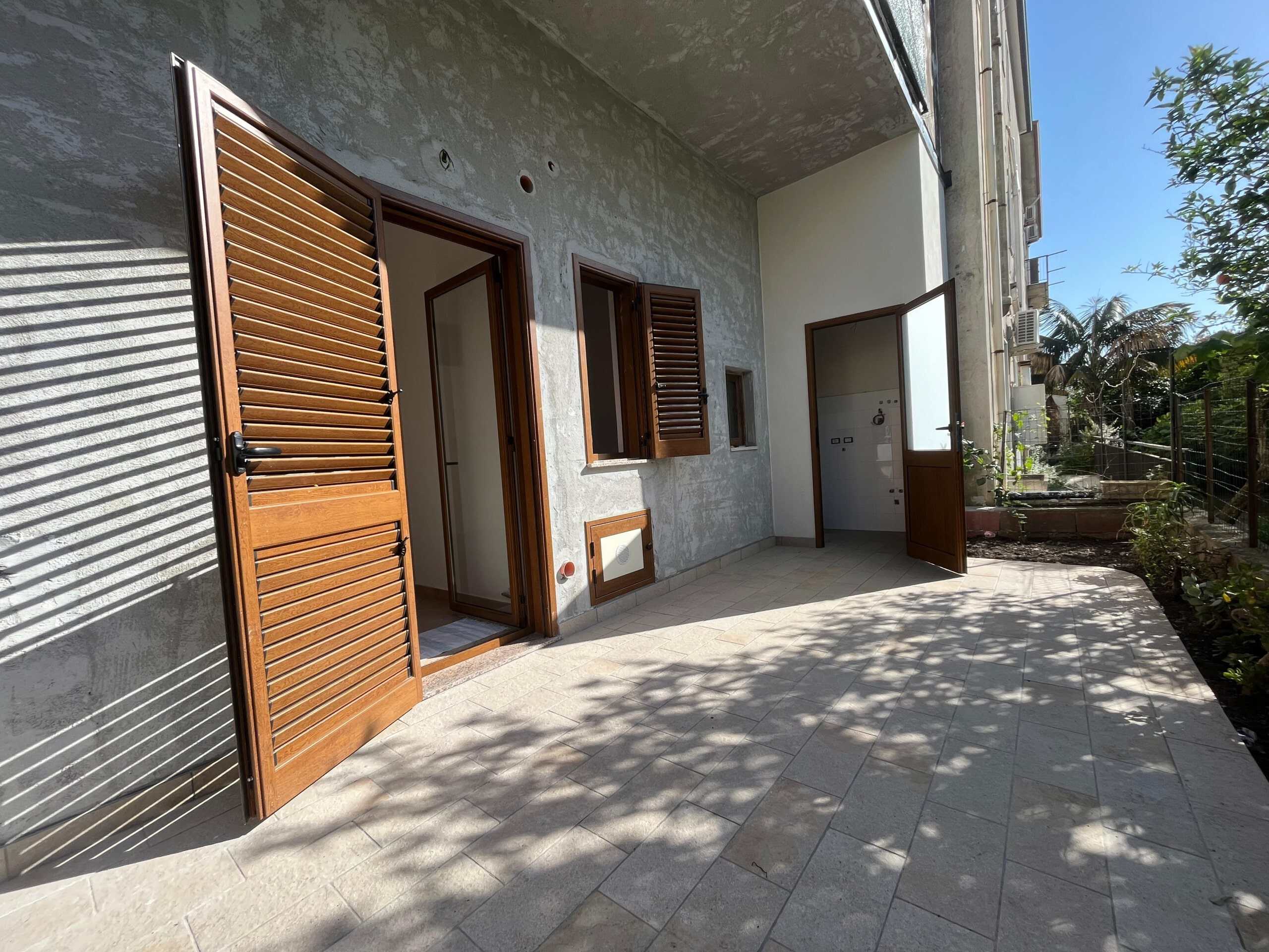 Finestra e porta in legno su terrazza esterna di un appartamento non finito, con pareti in calcestruzzo e pavimento in piastrelle, in un ambiente urbano soleggiato.