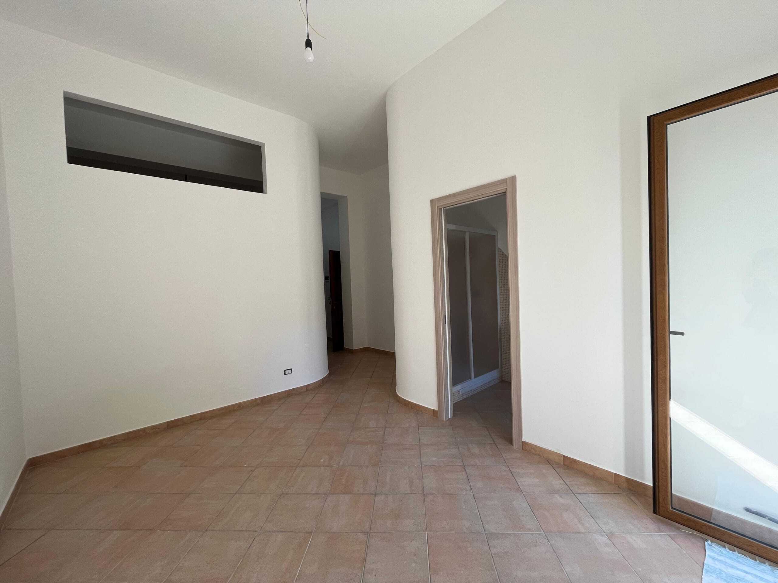 Ampio interno di un appartamento residenziale con pareti bianche, porta a vetri con cornice in legno, pavimento in piastrelle beige e grande finestra a soffitto, perfetto per vendita immobiliare.