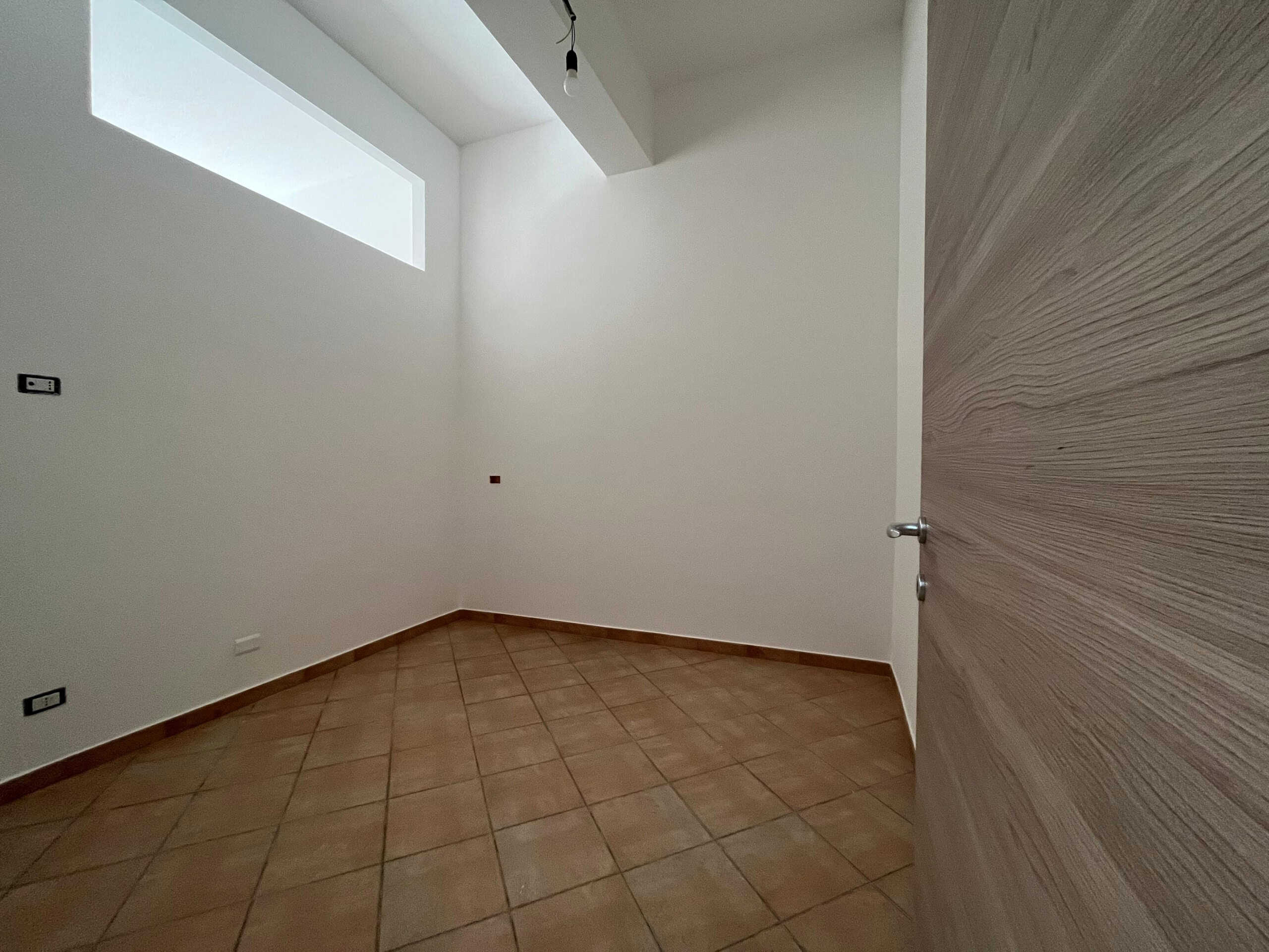 Spazio vuoto in appartamento con pareti bianche e pavimento in gres beige, finestra alta naturale, porta in legno chiaro, nuovo, pronto per arredare, immobile residenziale, NextCasa.