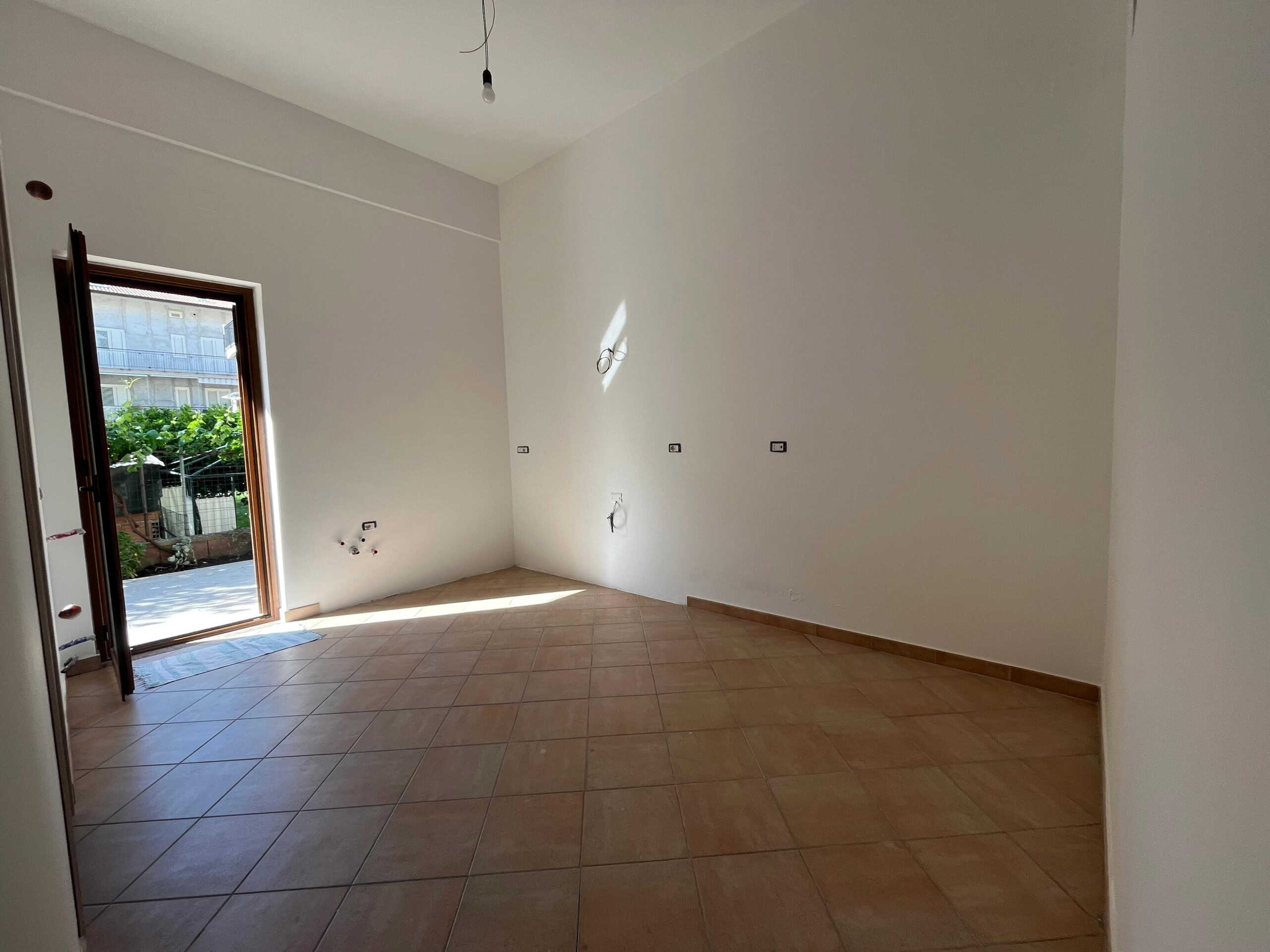 Spazio interno di una casa in fase di ristrutturazione con pareti bianche, pavimento in gres effetto terracotta e porta finestra che dà accesso a un balcone con vista verdeggiante.