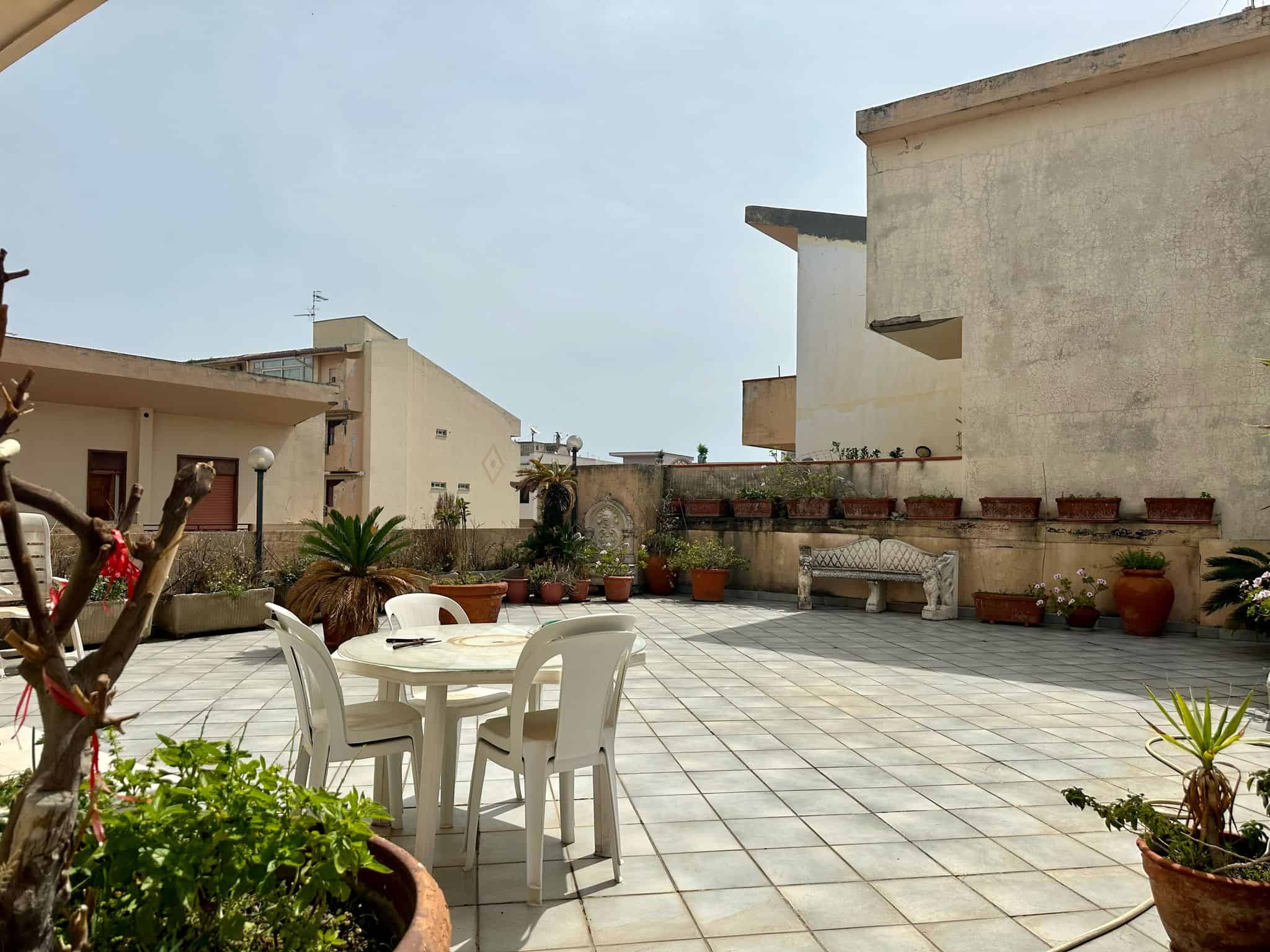Ampio terrazzo con piante in vaso, tavolo e sedie in plastica, ideale per relax e socialità, con vista sul cielo e sull'arredamento urbano, perfetto per vivere all'aperto in città.