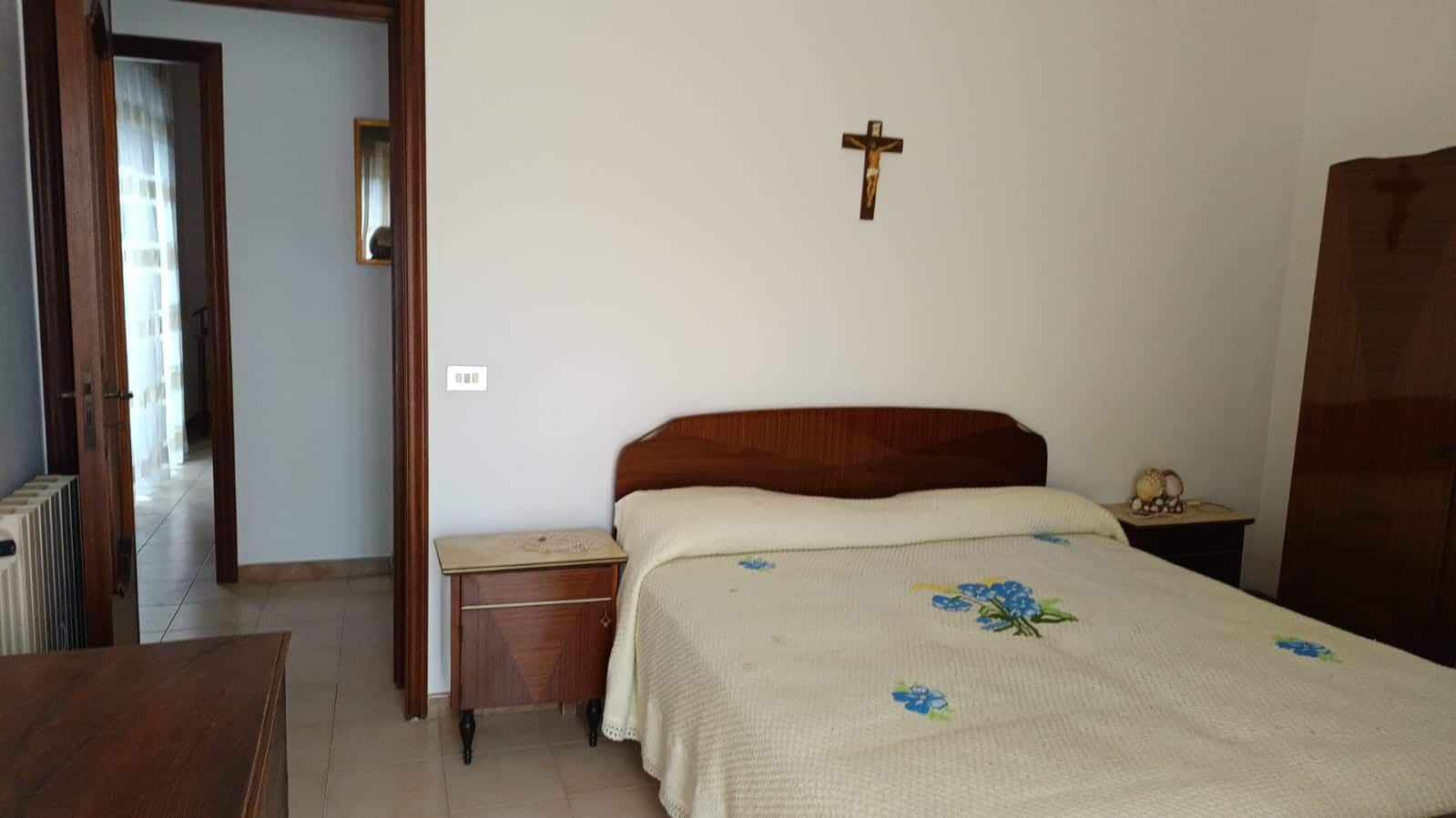 Letto matrimoniale con arredamento classico in legno, camera da letto luminosa con porta verso il corridoio, parete con croce in legno, ambienti accoglienti e funzionali.