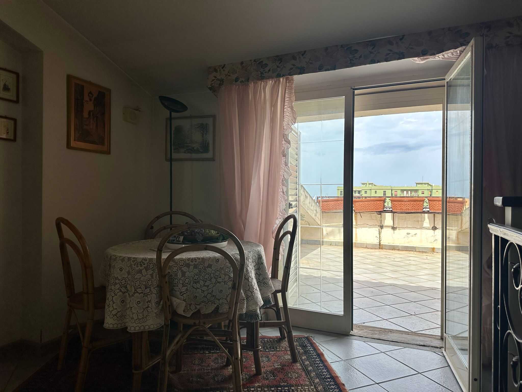 Vista interna di una sala da pranzo con tavolo tondo ricoperto da tovaglia in pizzo e sedie in legno, aperta su un ampio balcone soleggiato con vista su edifici moderni, luce naturale, atmosfera accogliente.