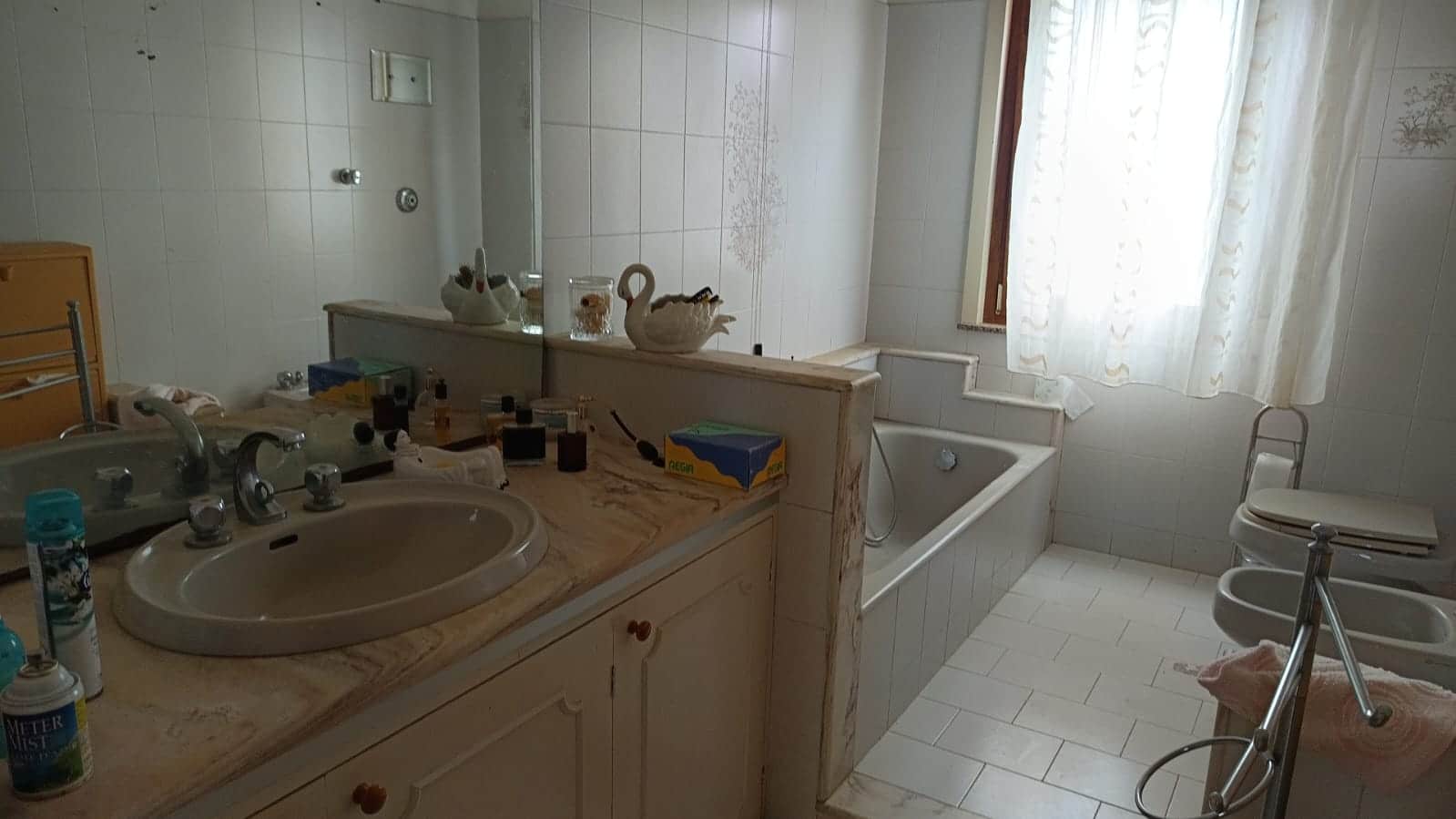 Fotografia di un bagno semplice con lavabo, vasca da bagno e bidet, decorato con statuette e accessori bagno, illuminato da luce naturale tramite finestra con tende chiare.