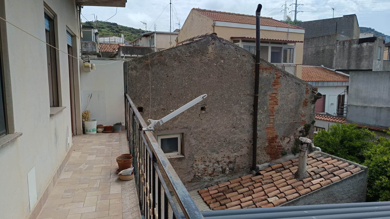 Balcone con tende da sole e piante in vaso, vista su tetti e le abitazioni vicine in un quartiere di città in collina, con un ambiente urbano caratteristico e un cielo nuvoloso.