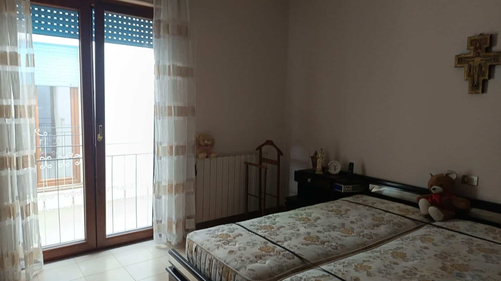 Att nafto potente e luminosa con balcone e tende trasparenti, ideale per ambienti residenziali. La stanza dispone di letto, decorazioni e accessori come teddy bear e croce religiosa. Perfetta per appartamenti in vendita o affitto.