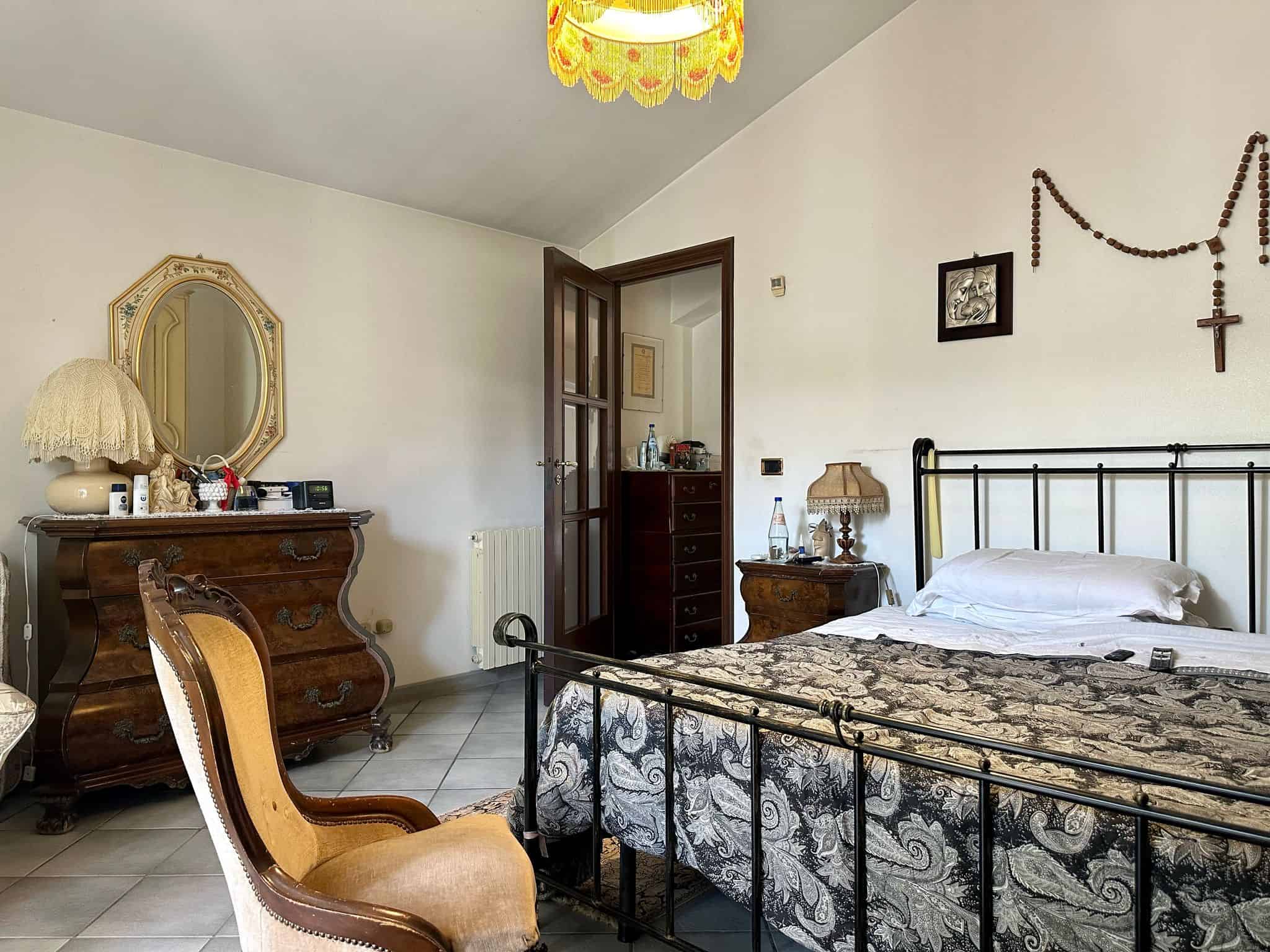 Comodino con lampada da tavolo, cassettiera in legno in stile classico, letto con testiera in metallo nero e copriletto a fantasie, decorazioni religiose sulla parete, arredamento vintage, dettaglio interno di camera da letto, atmosfera accogliente.