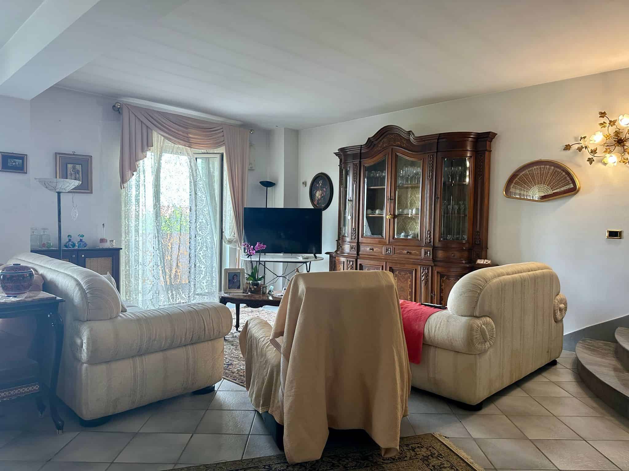 Ampia living room con arredi classici, divani beige, grande mobile antico in legno, TV e decorazioni eleganti, ambiente luminoso con finestre a volontà, ideale per il comfort domestico.