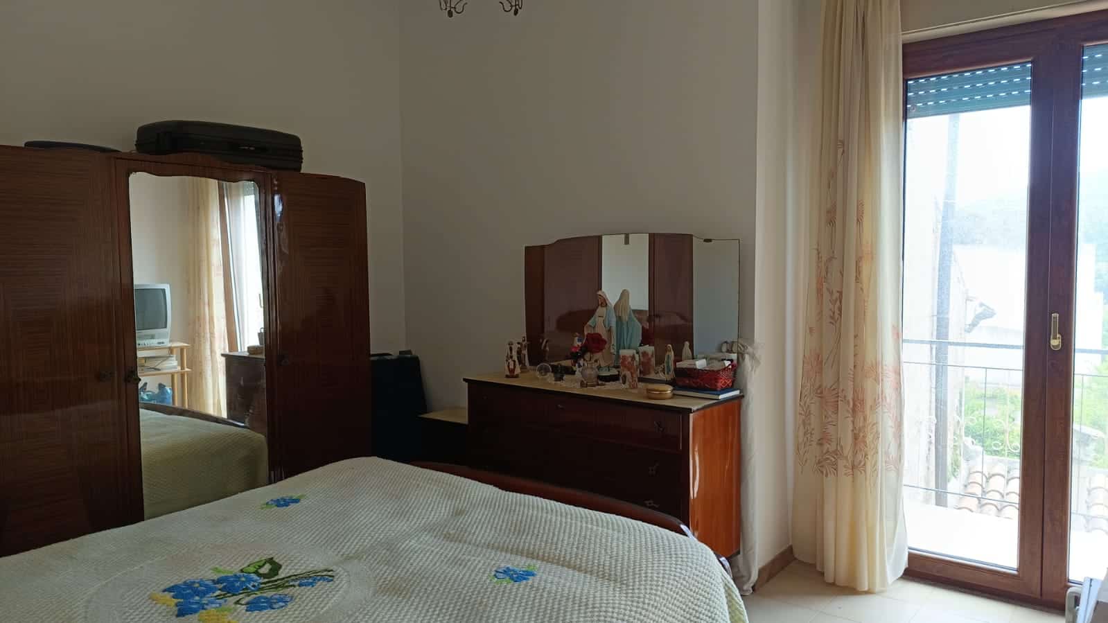 Armadio in legno con specchio e mobile con decorazioni religiose in una camera da letto, con porta finestra che conduce a un balcone, arredamento classico e accogliente.