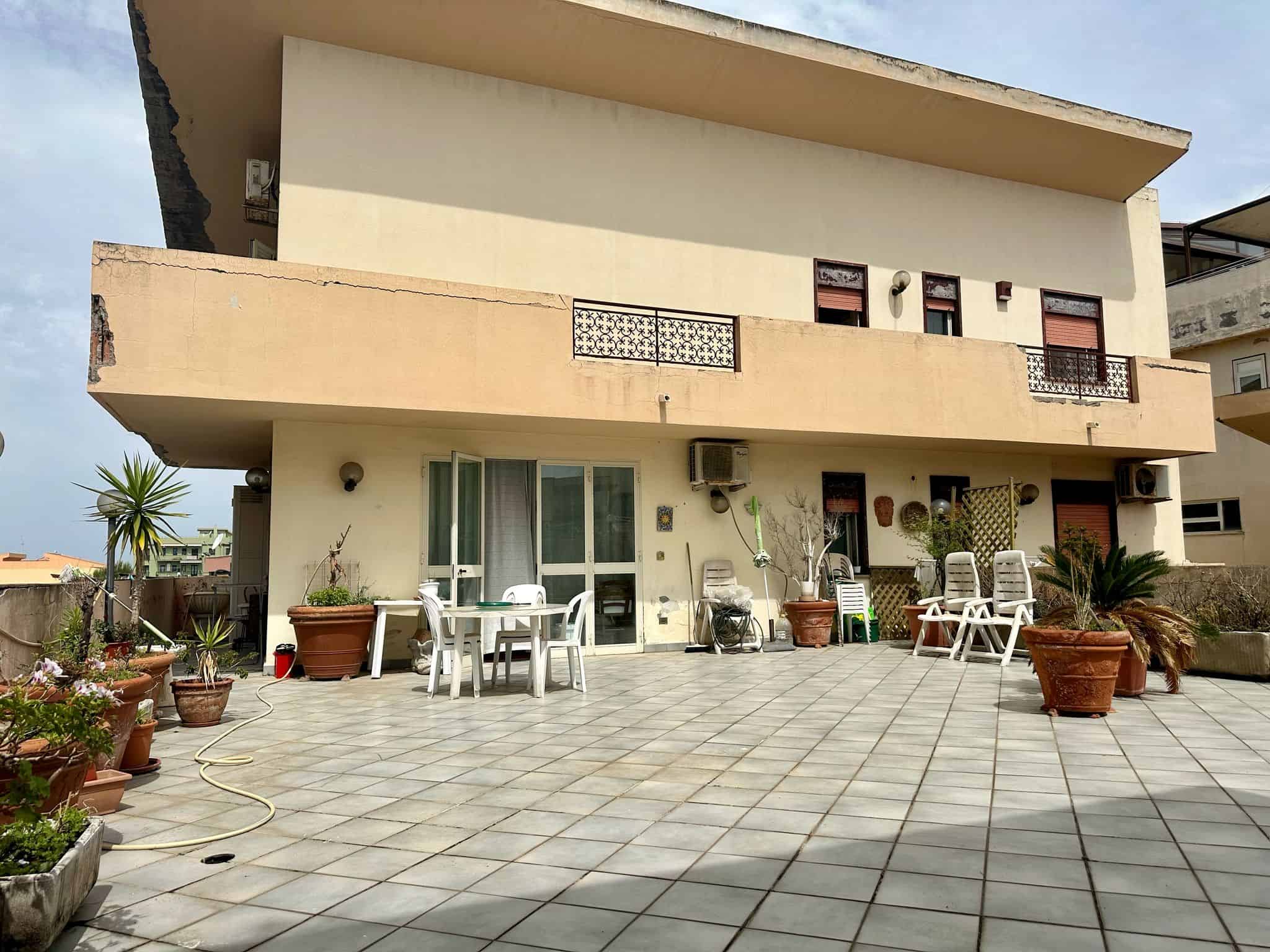 Ampio terrazzo con decorazioni floreali, piante in vaso e mobili da esterno, vista su edificio residenziale, ideale per relax e socializzazione.