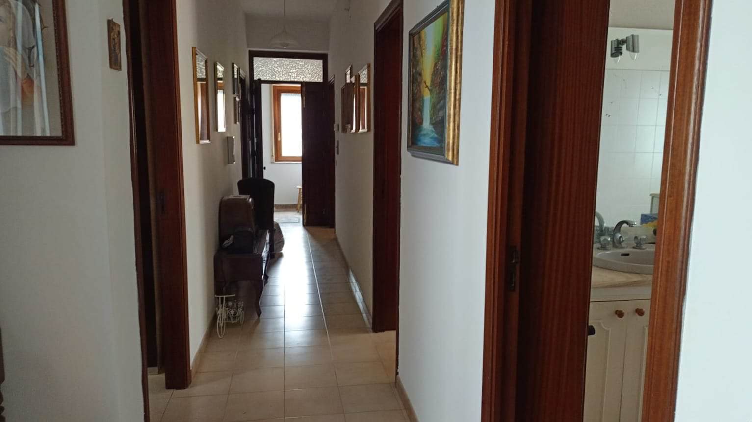 Bagno di un appartamento con pareti chiare, porte in legno e dipinti alle pareti, corridoio con parquet chiaro e finestra luminosa, stile classico e accogliente.