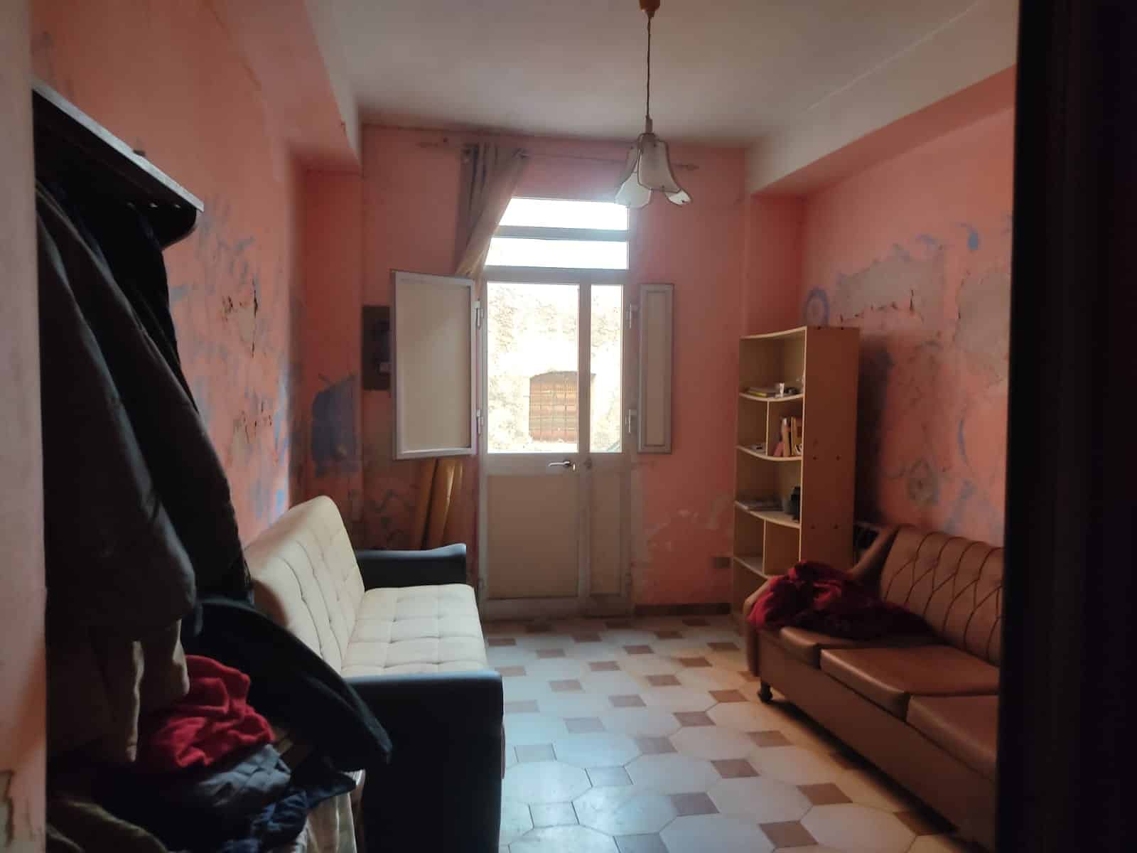 Soggiorno con pareti color pesca, divani e libreria, finestra aperta con tenda chiara, ambiente luminoso e accogliente, arredamento semplice, pavimento in piastrelle a scacchiera.