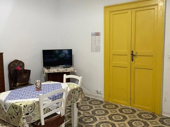 Soggiorno con pareti bianche, ingresso con porta gialla, tavolo con tovaglia floreale e sedie in legno, TV su mobile, pavimentazione in piastrelle decorative, elementi d'arredo tradizionali.