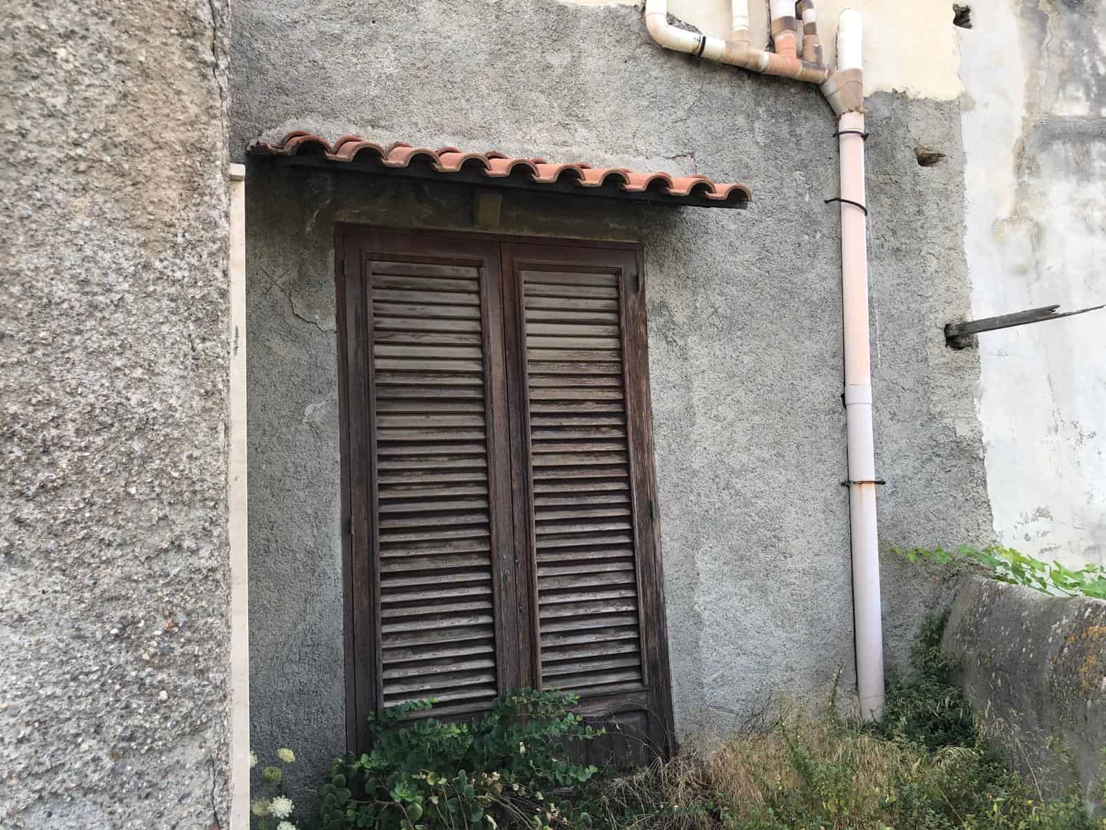 Finestra in legno con persiane arrugginite su parete in pietra, stile rustico, contesto urbano con dettagli di tubature e vegetazione sottostante.