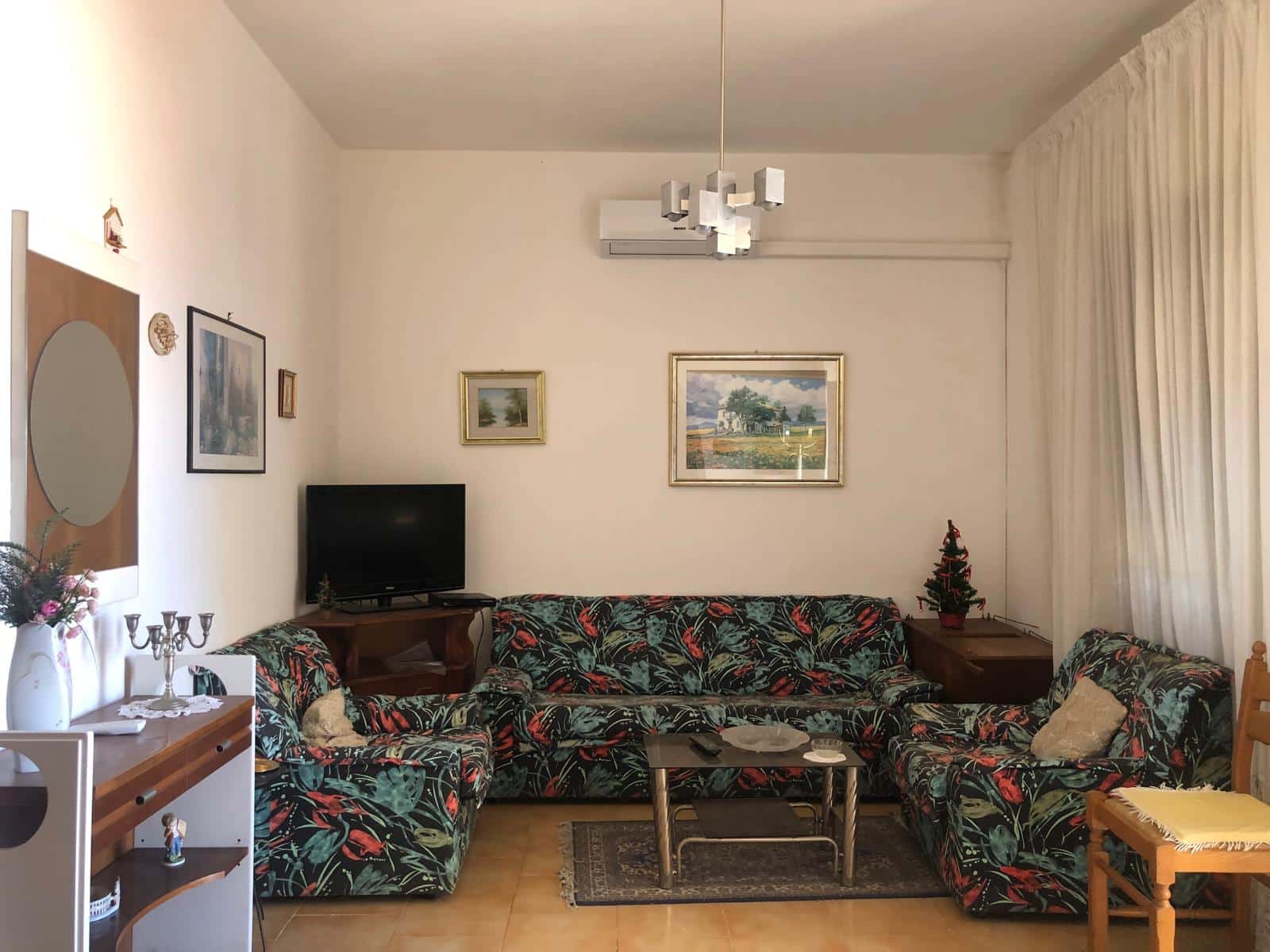 Sofà con rivestimento a motivo tropicale in soggiorno, arredamento accogliente con quadro paesaggistico, televisore e decorazioni natalizie, atmosfera calda e familiare, casa NextCasa in Italia.