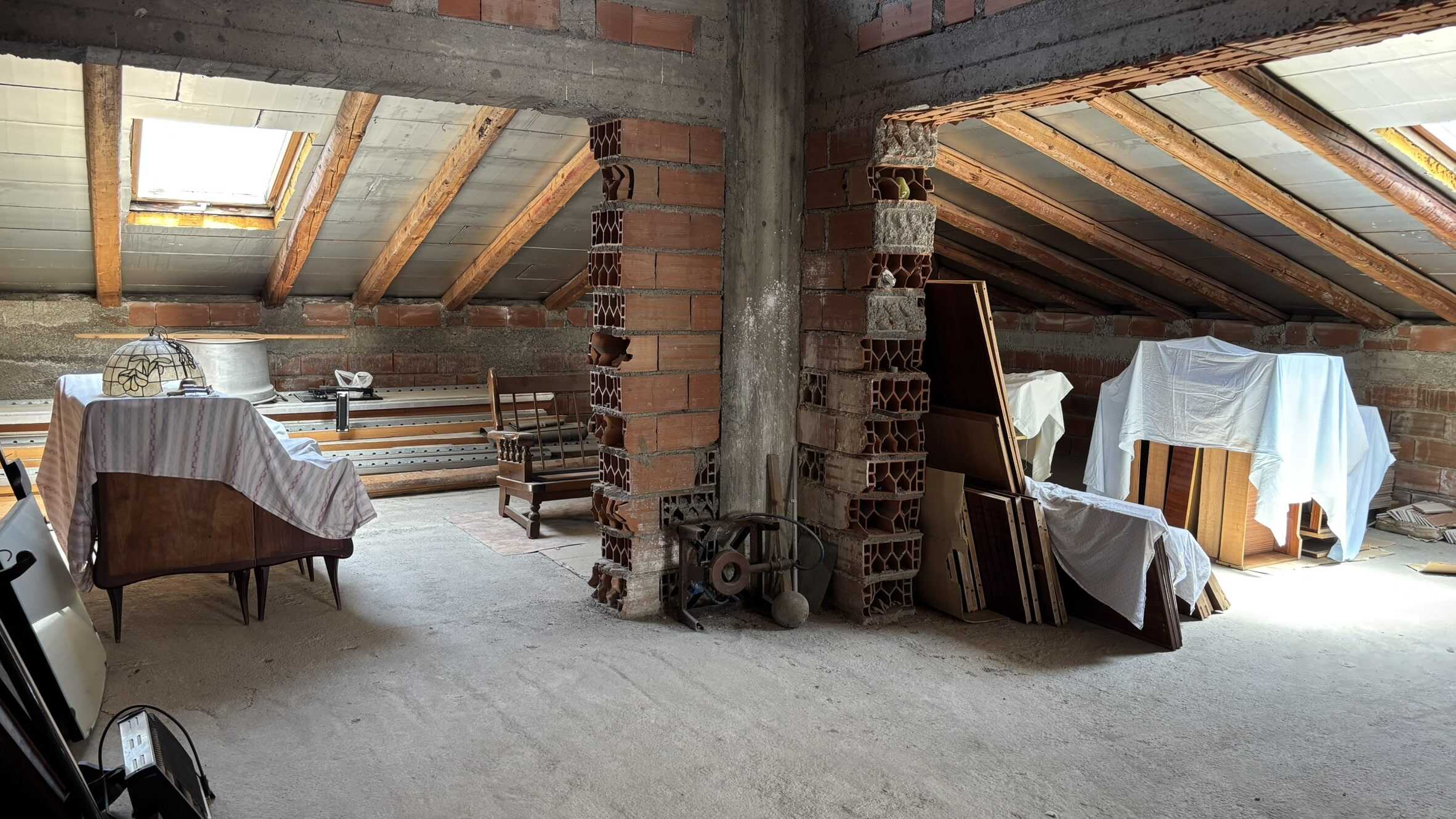 Appartamento in fase di ristrutturazione con travi in legno e finestre Velux per la luminosità naturale. Spazio attico con pareti in mattoni e arredi parzialmente coperti, ideale per progetti di interior design.