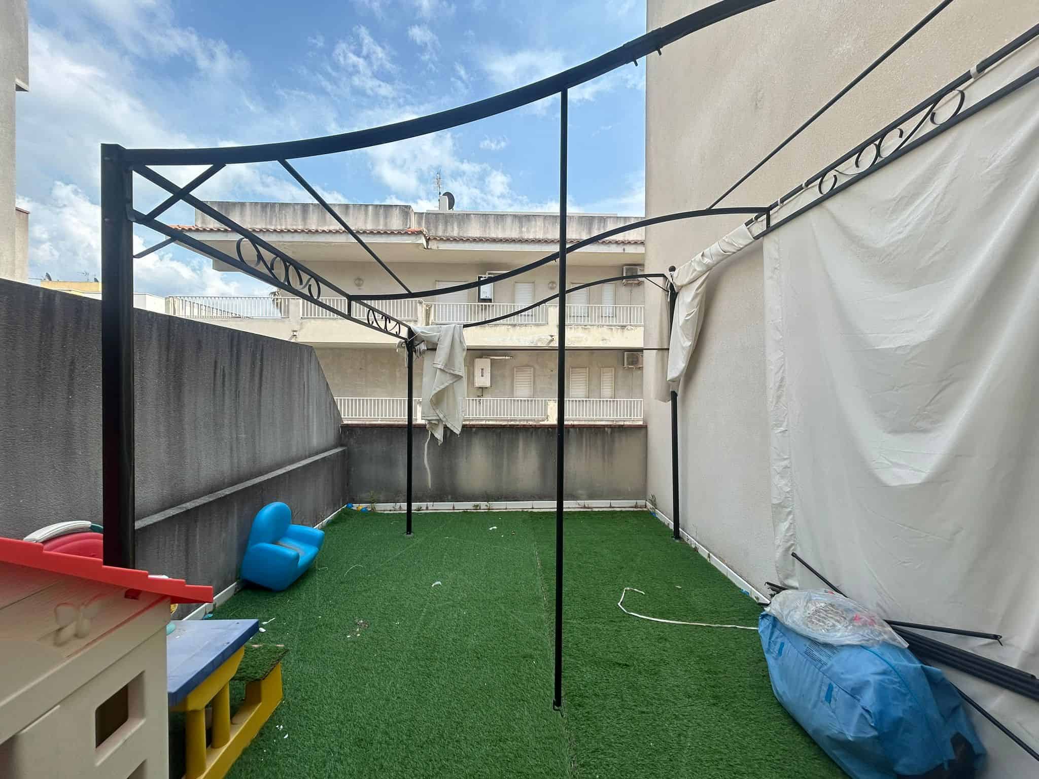 Balcone di appartamento con tappeto erboso artificiale, spazio esterno con filo per bucato, arredi per bambini e vista su edifici residenziali in città, ideale per relax e attività all'aperto.