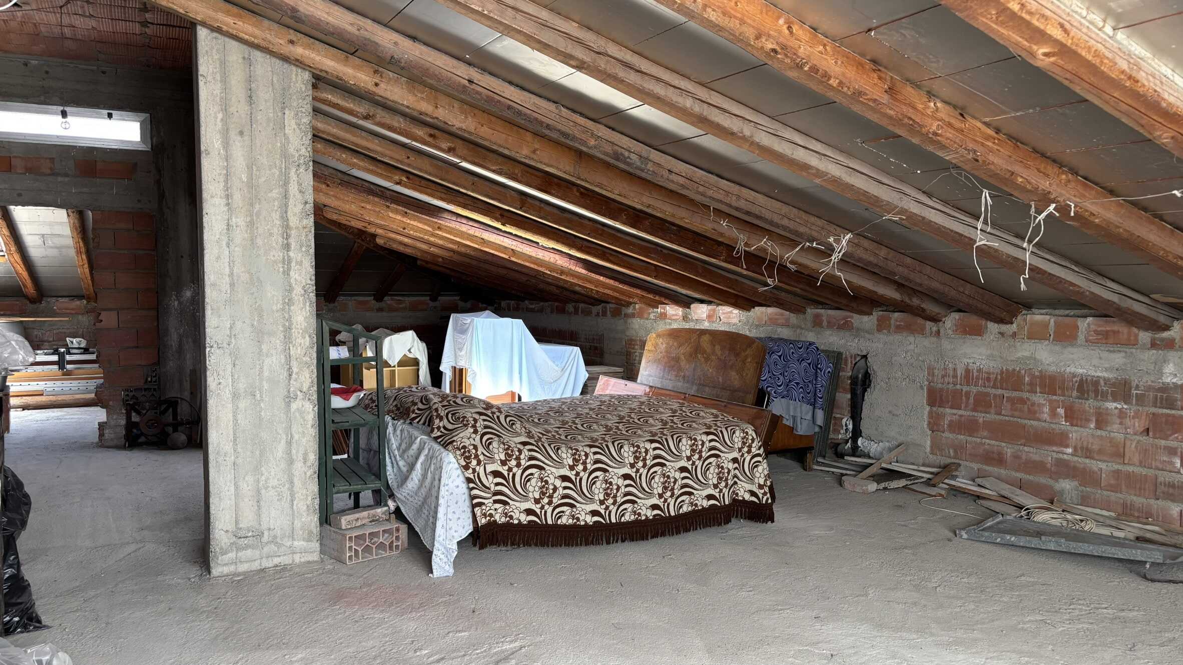 Vecchio sottotetto grezzo in fase di ristrutturazione, con travi in legno e pareti in mattoni, arredato con vecchi mobili e stoffe di copertura. Ideale come spazio per un progetto di restauro o un loft alternativamente ristrutturato.
