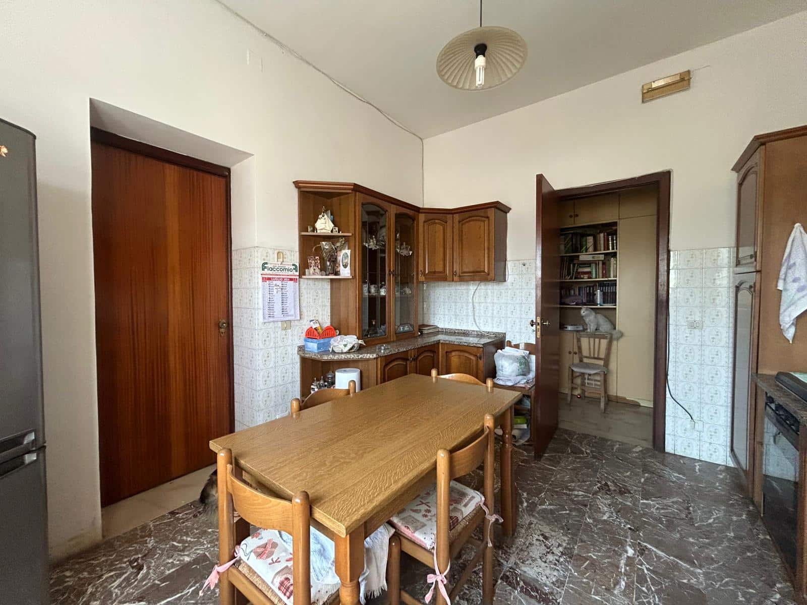 Vecchia cucina con mobili in legno, tavolo da pranzo e pavimento in marmo, pareti rivestite con piastrelle beige, ambiente domestico arredato in stile tradizionale.