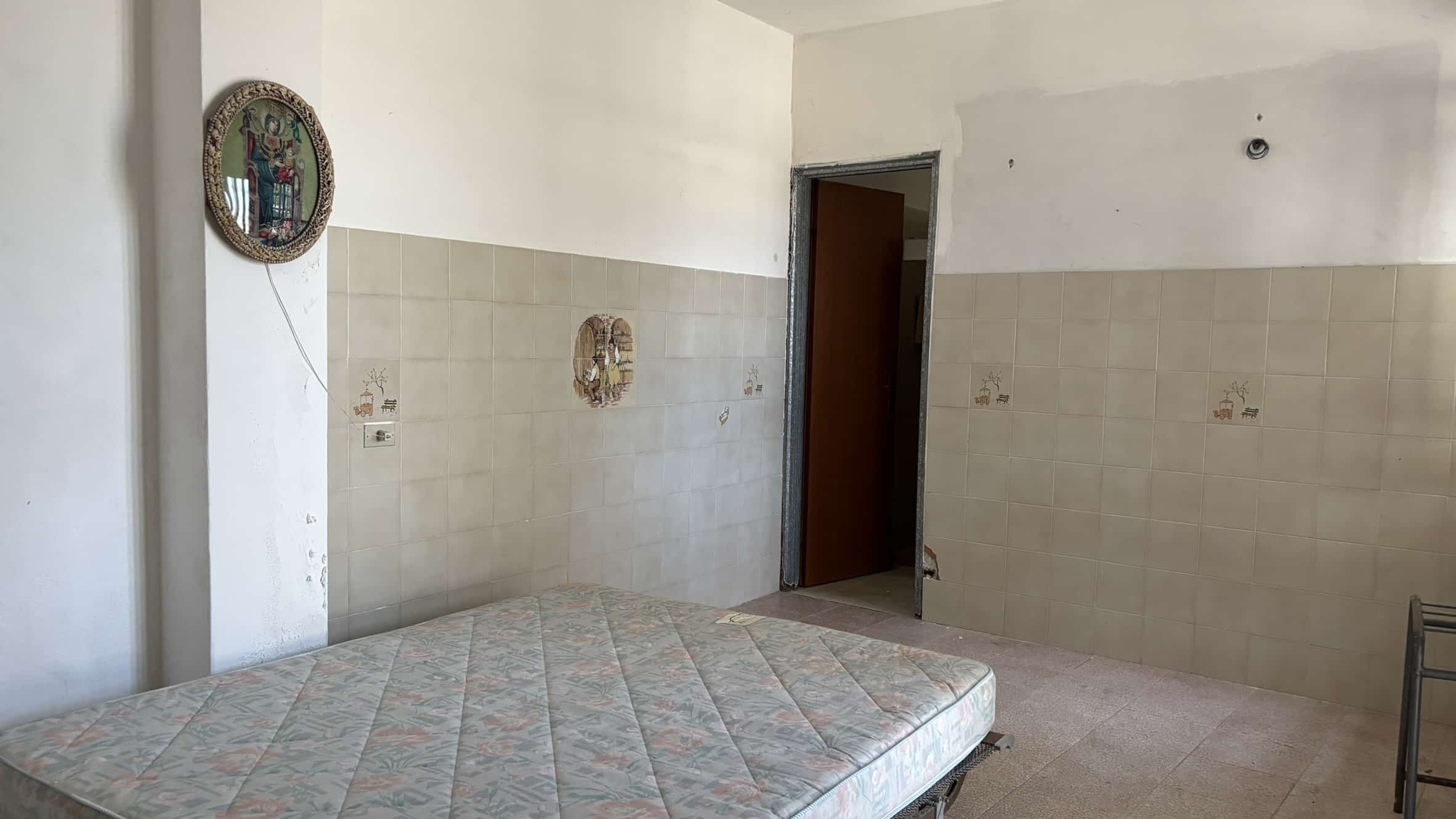 Letto singolo con materasso in una stanza in fase di ristrutturazione, pareti con piastrelle decorative e scena religiosa, ambiente semplice e funzionale, ideale per affitti o ristrutturazioni immobiliari.
