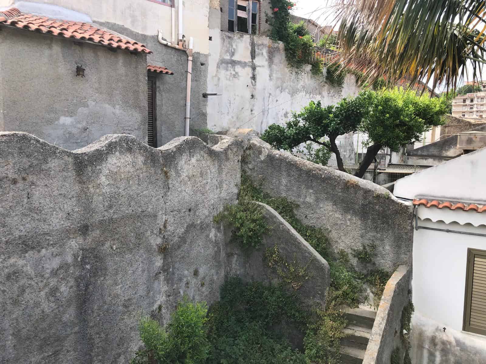 Vecchio muro in pietra con piante ornamentali e scale in pietra, ambiente urbano con elementi storici e vegetazione, cortile interno di edificio residenziale in Italia.