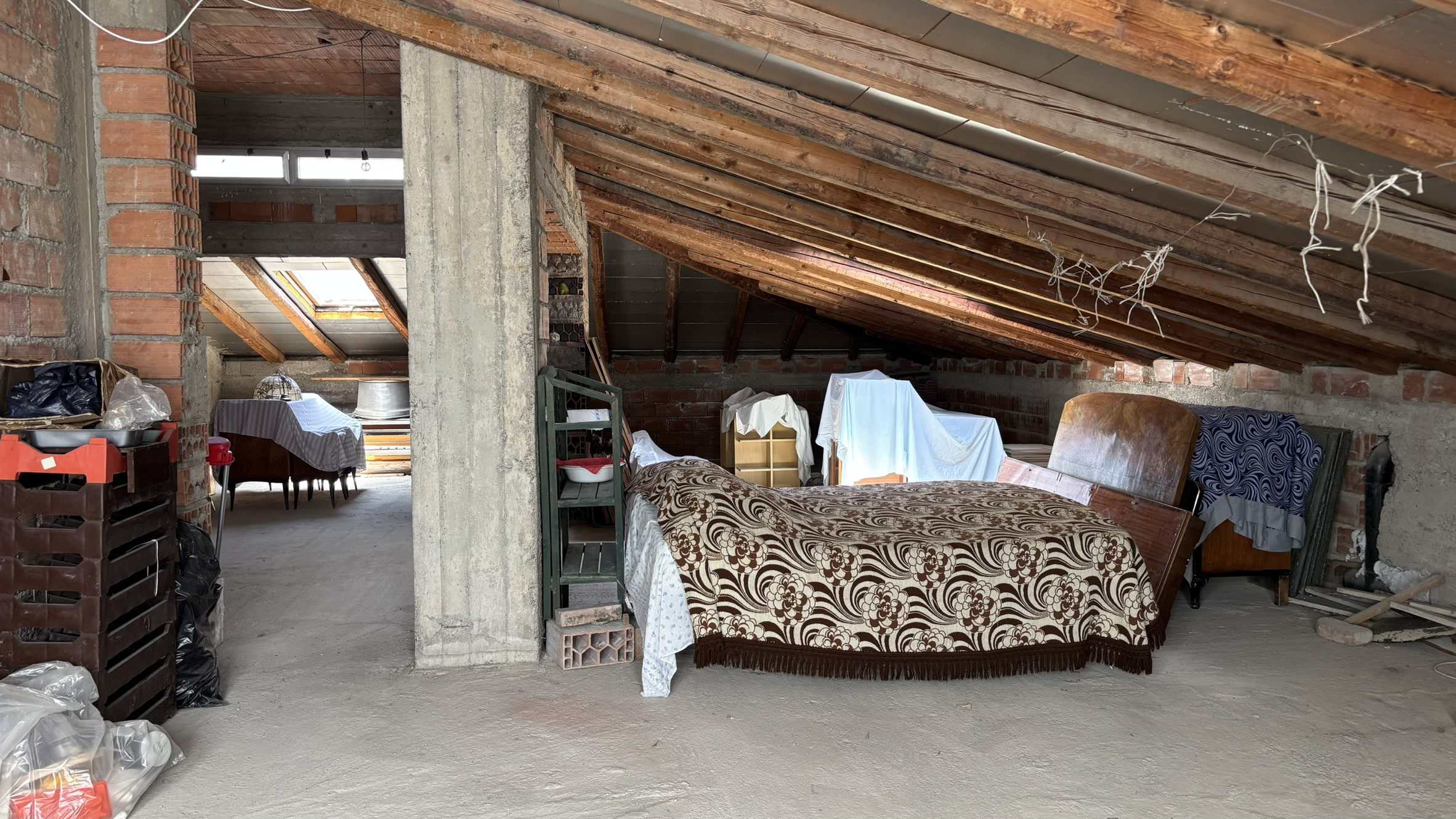 Letto in mansarda in fase di ristrutturazione, ambientazione rustica con tetto in legno e mattoni a vista, materiali da costruzione e arredi provvisionali, spazio abitativo in fase di completamento, casa di design NextCasa, soluzione immobiliare innovativa.