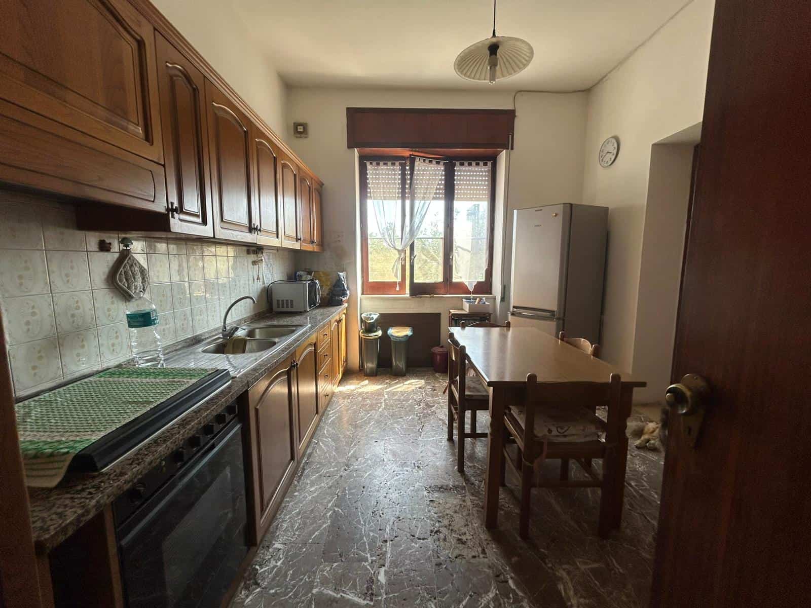 Cucina abitabile con ante in legno, piano in marmo, lavello doppio, frigorifero, tavolo da pranzo e sedie, finestra grande con tende, pavimentazione in marmo, elettrodomestici e arredamento classico.