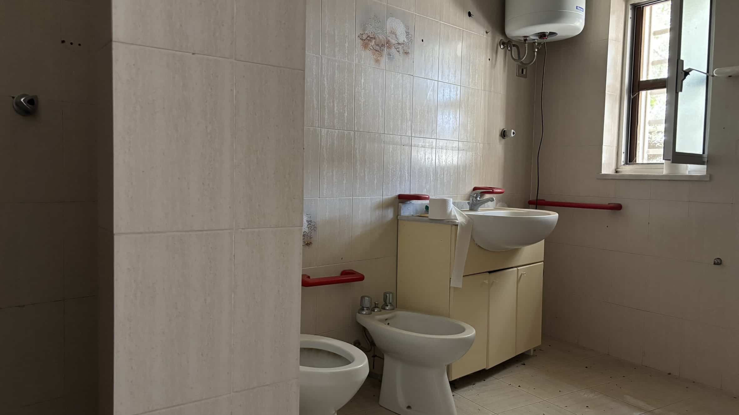 Bagno con lavabo, bidet e water in ambiente spazioso e luminoso, con piastrelle beige e finestra che permette l'ingresso di luce naturale, ideale per servizi igienici funzionali e moderni.