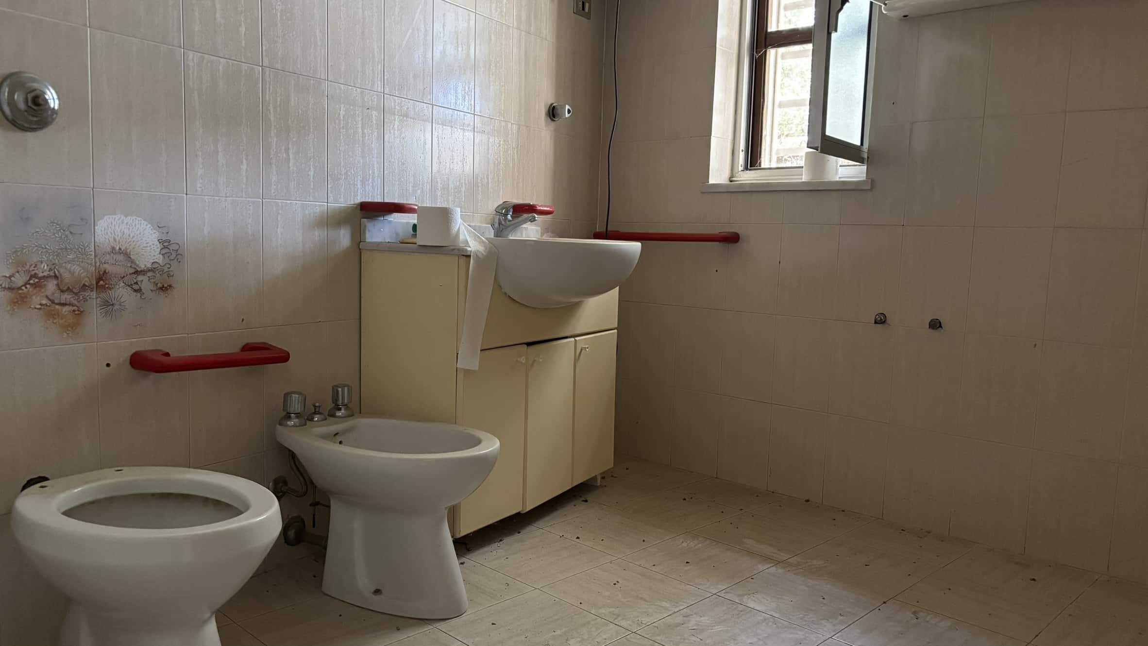 Lavabo e bidet in bagno con pareti e pavimenti in piastrelle beige, bagno in fase di ristrutturazione, finestra aperta, accessori di sicurezza per disabili, spazio vuoto attorno alle utenze.
