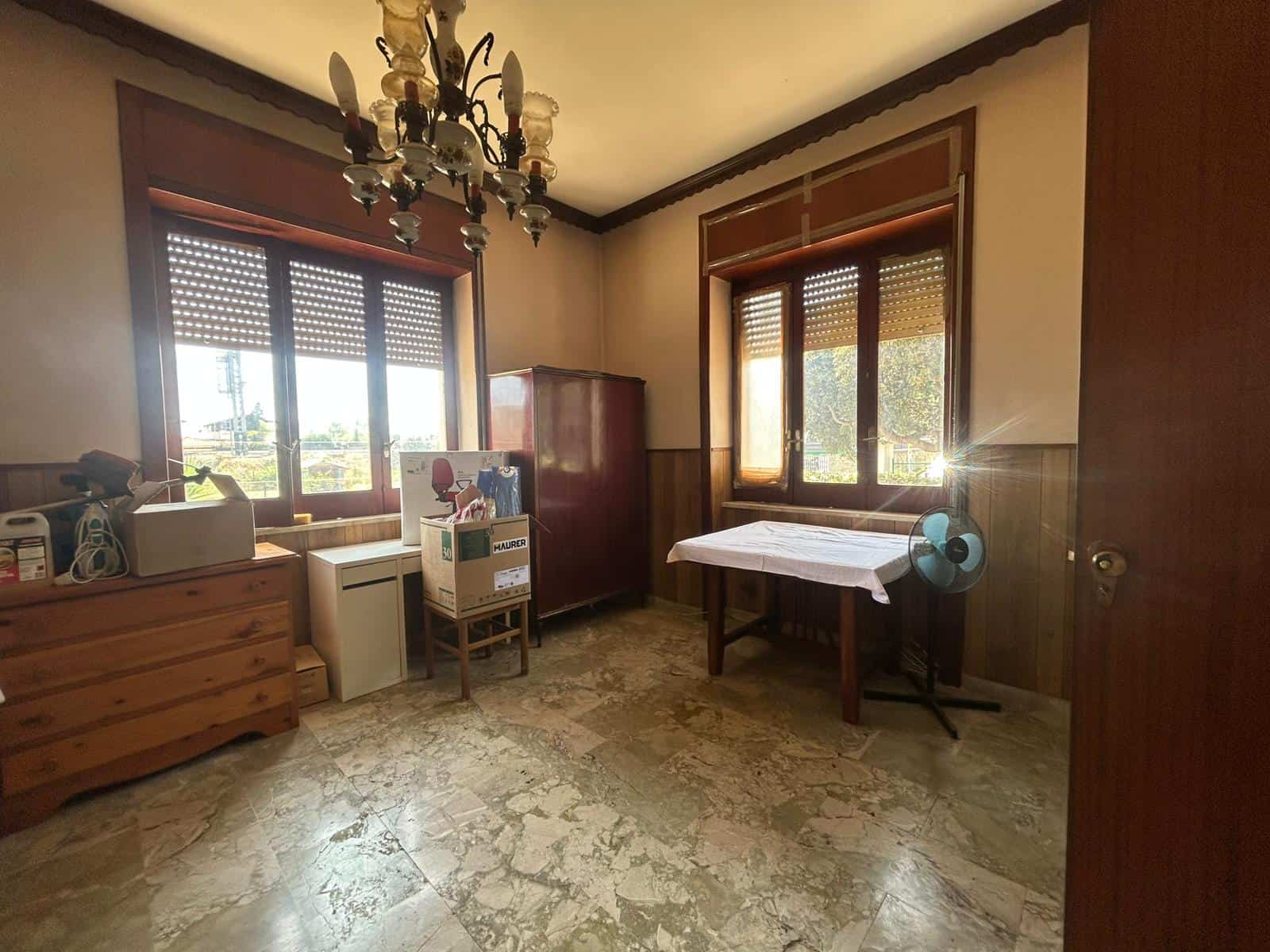 Stanza spaziosa con grandi finestre, pavimento in marmo, arredi in legno, lampadario anticato, ideale come ufficio o soggiorno, esposta al sole con luce naturale diffusa, stile classico e accogliente.