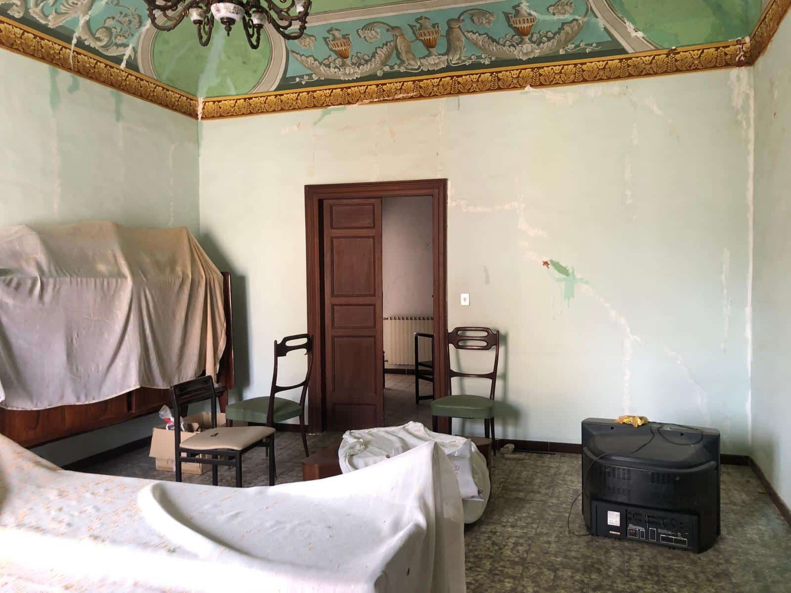 Antico interno abitazione con soffitto affrescato, pareti in fase di restauro, mobili d'epoca, televisore vintage e sedie ancien style, ambiente storico da ristrutturare.