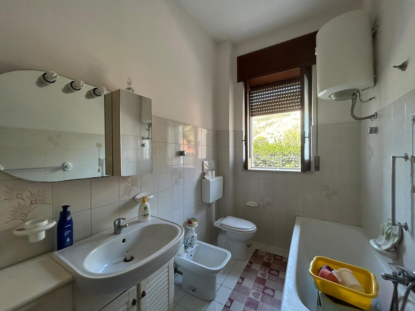 Lavabo e servizi igienici in un bagno di casa, con finestra che permette l'ingresso di luce naturale, pareti rivestite di piastrelle beige con decori floreali, e accessori per la cura personale.