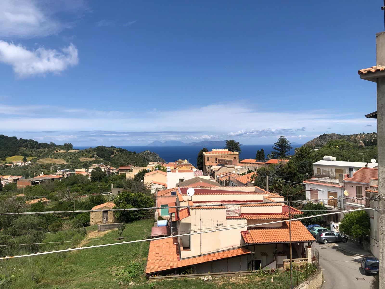 Paesaggio urbano mediterraneo con case e tetti in terracotta, vista mare e colline verdi, cielo azzurro e nuvole, eccezionale panorama in campagna, residenza tranquilla vicino al mare, immobiliare NextCasa Italia.