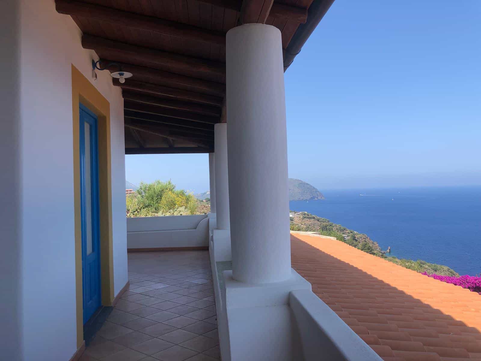 Vista panoramica mare dalla terrazza di una casa in stile mediterraneo, con colonne bianche, tetto in legno e panorama di coste e isole, ideale per case di lusso in affitto o vendita.