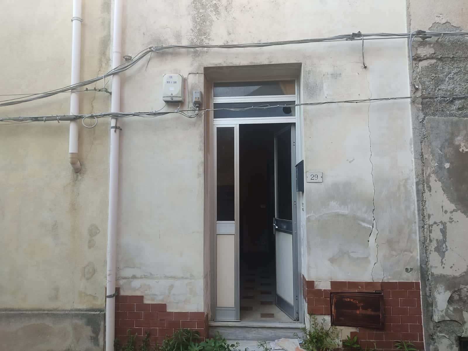 Vecchio ingresso di una casa con porta in metallo e pareti con crepe e segni di usura, dettagli di tubature visibili, in zona con decorazioni di piastrelle rosse e piccoli piante sul pavimento.