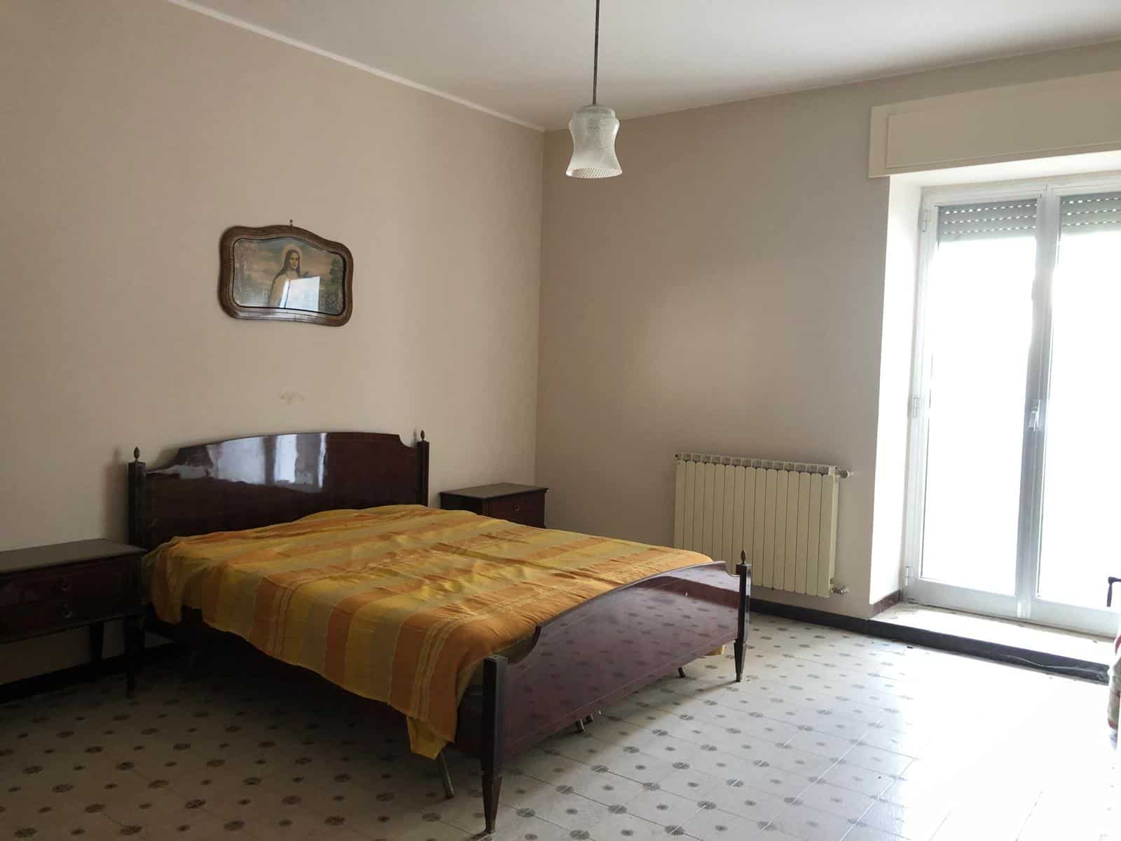 Letto matrimoniale in camera da letto, arredamento classico, finestra grande con balcone, ambiente luminoso, riscaldamento centrale, pavimento in piastrelle, decorazioni semplici, stile anni passati, soluzione abitativa confortevole, casa da affittare o vendere, immobile residenziale.