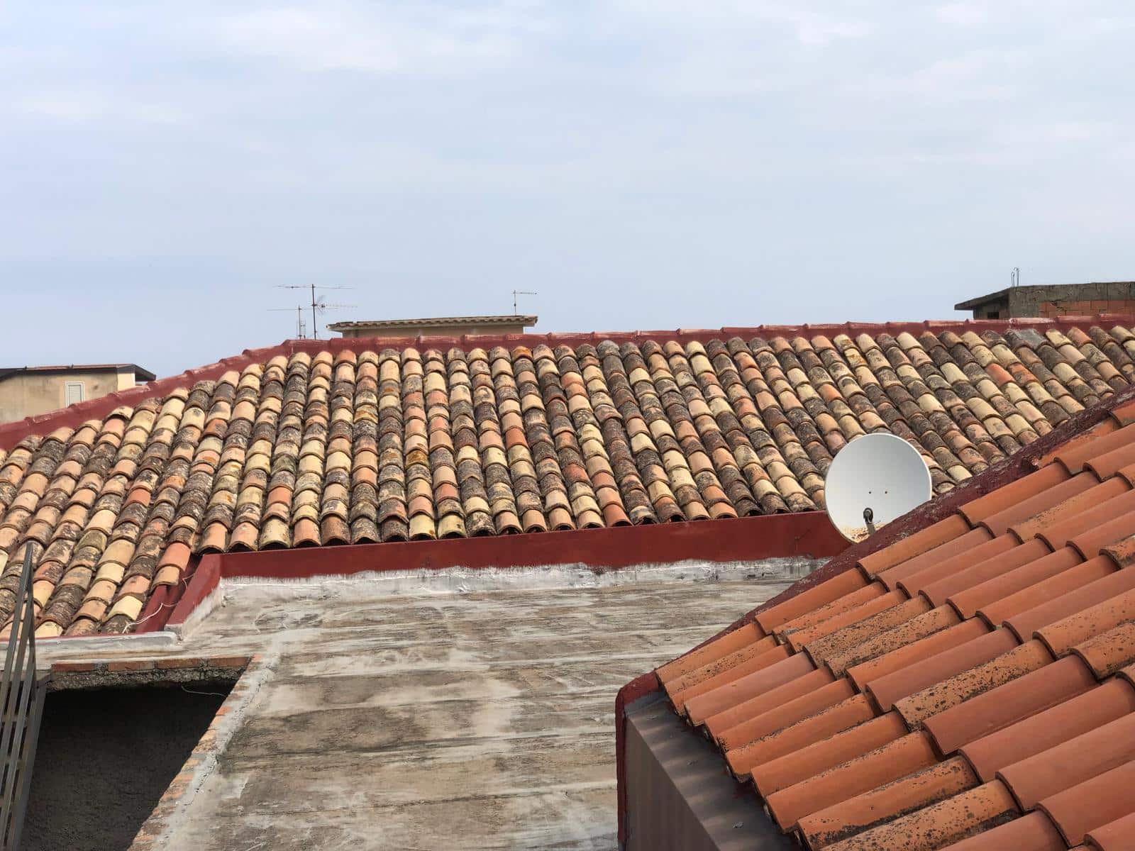 Tegole di terracotta rossa e smaltata su tetto di case in città, con antenne TV sullo sfondo e un cielo nuvoloso, rappresentando l'architettura residenziale tradizionale italiana.