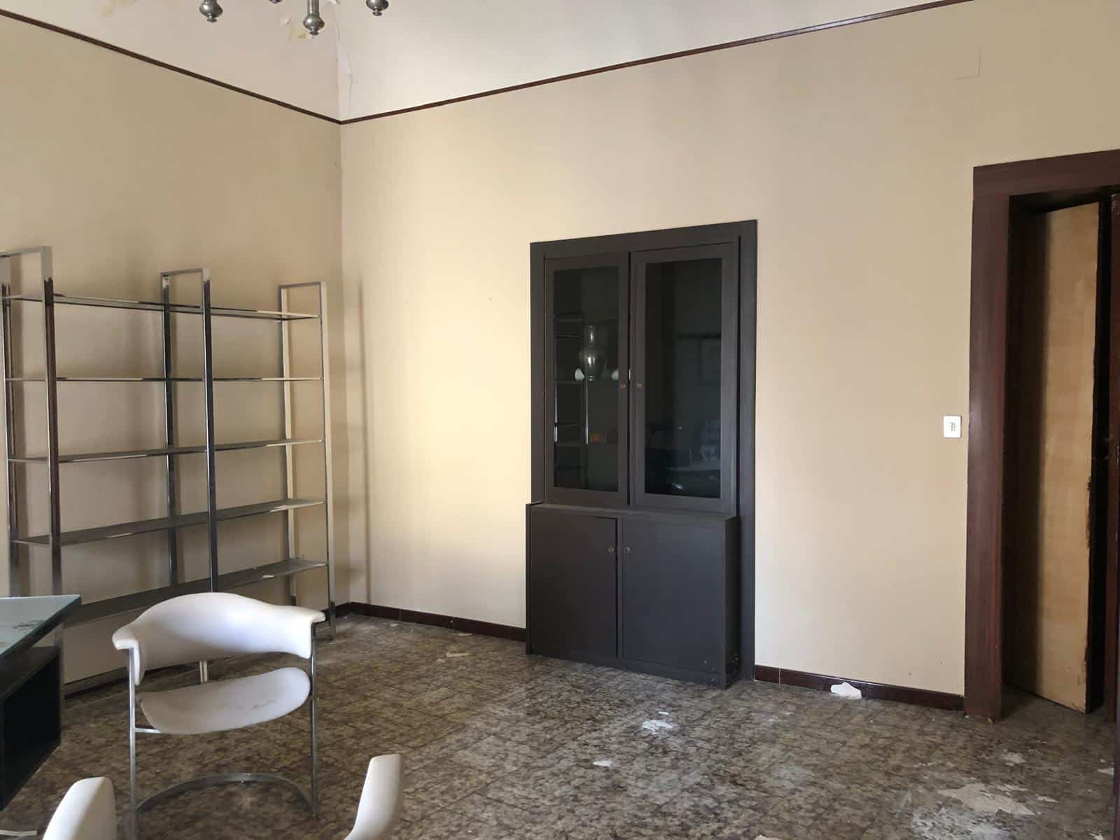L'immagine mostra un soggiorno con un arredamento semplice e funzionale, caratterizzato da una libreria moderna in metallo, una credenza in legno scuro con ante in vetro e una cornice in legno intorno alla porta. La stanza ha pareti di colore beige e un pavimento in marmo a scacchi, creando un ambiente pratico e sobrio, ideale per soggiorni o aree di lavoro domestiche. La disposizione consente un facile accesso agli spazi e permette di personalizzare l'ambiente secondo le esigenze.