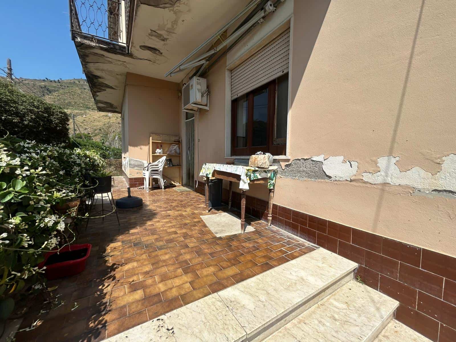 Balcone di casa con pavimento in ceramica e piante, vista sul paesaggio circostante con colline e vegetazione, spazio esterno ideale per relax e conviviale, abitazione con pareti esterne in condizioni da ristrutturare, esposizione al sole.