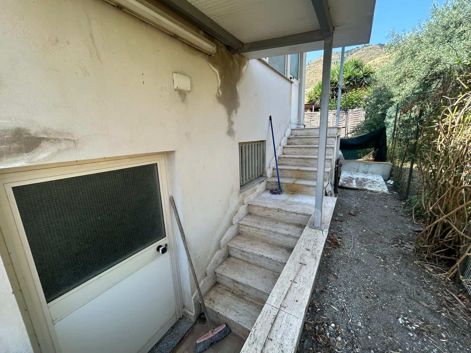 Esigenza di ristrutturazione esterna di casa in stile mediterraneo, con scale in pietra e ingresso al piano terra, l'area esterna necessita di pulizia e manutenzione.