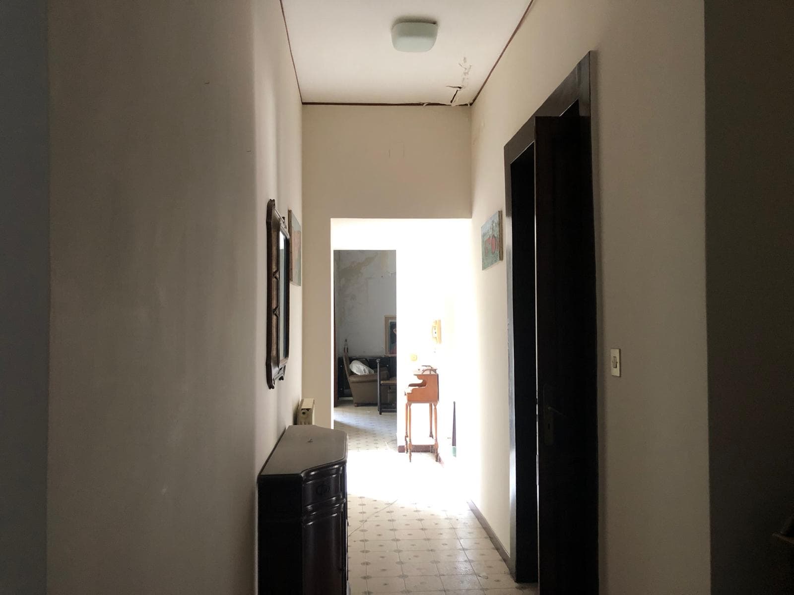 Vista di un corridoio di casa con pareti chiare, pavimento in piastrelle, dipinti alle pareti e porta scorrevole in legno, che conduce a una zona soggiorno luminosa e accogliente.