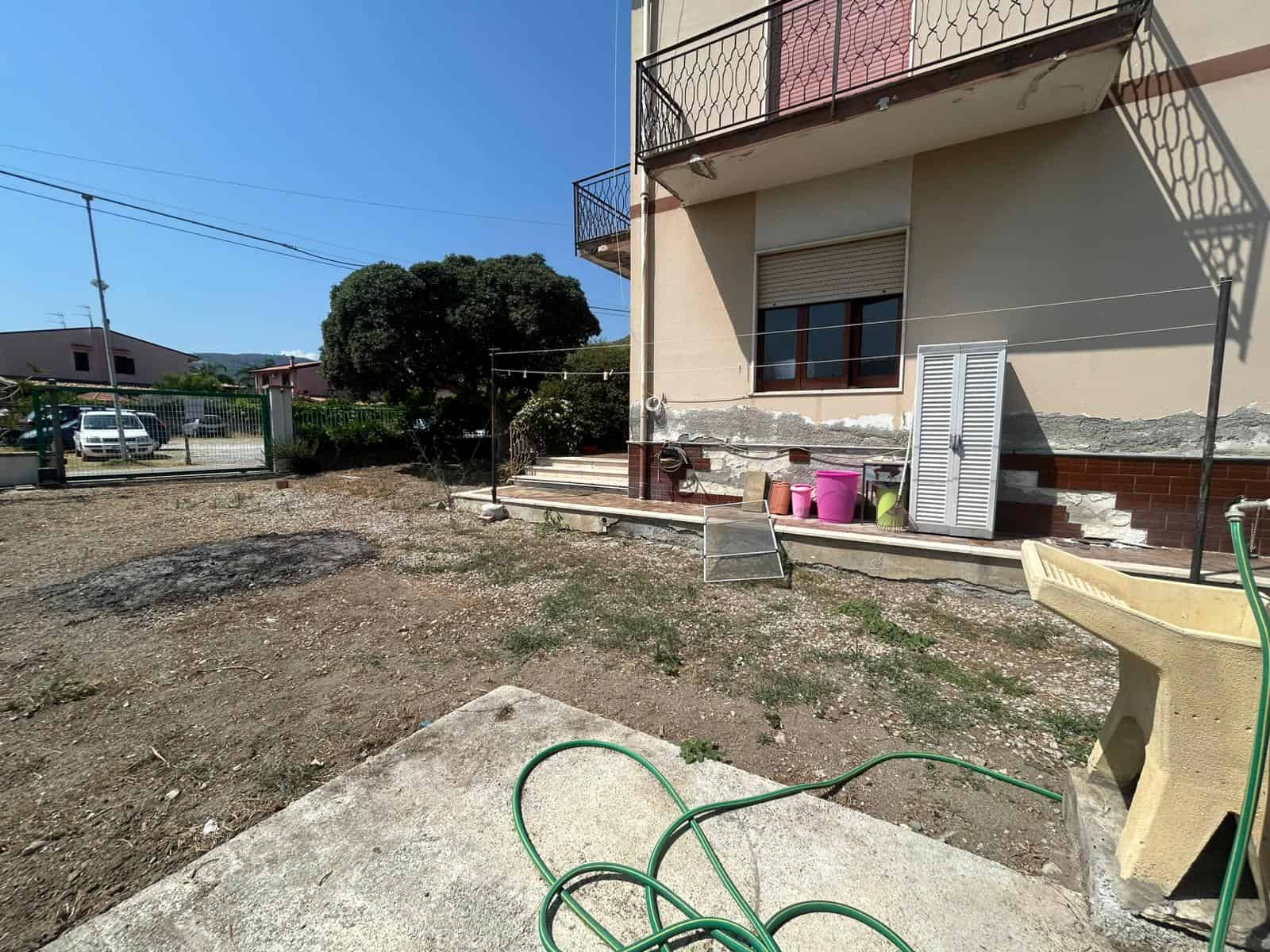 Veranda esterna di una casa in fase di ristrutturazione, con pavimentazione in cemento, attrezzi e bottiglie di plastica, vicino a un cancello in ferro che delimita il giardino.