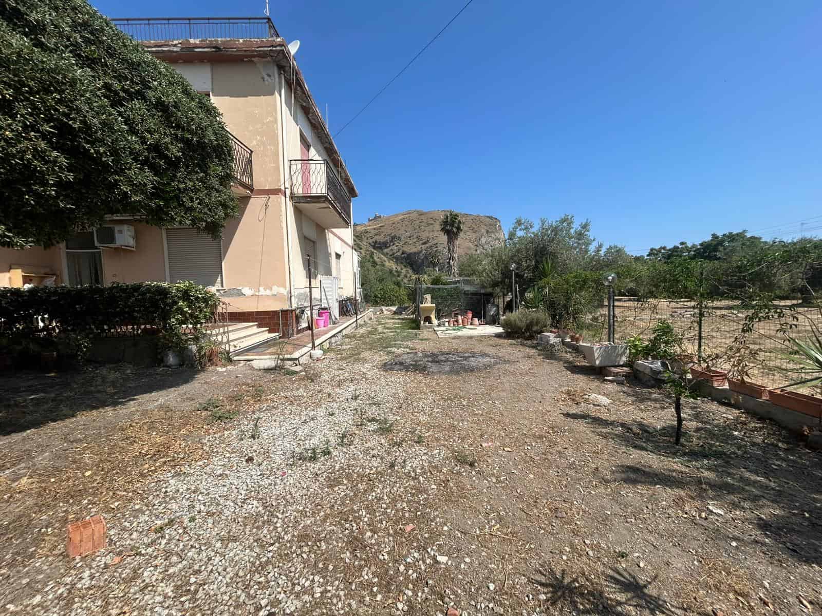 Zona esterna di una casa con giardino e terreno arido, vista sulla collina e cielo blu, ideale per immobili residenziali e investimento immobiliare.