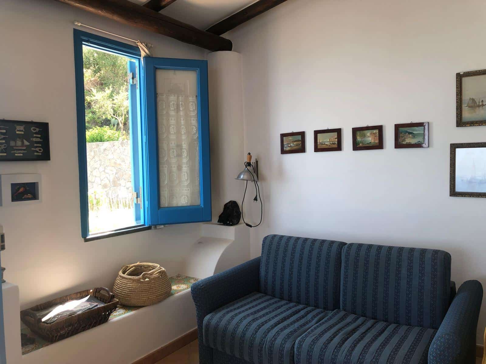 Finestra blu aperta in una stanza con decorazioni mediterranee, arredamento in stile classico, seduta in tessuto blu, pareti bianche con quadri e oggetti d'antiquariato, atmosfera accogliente e luminosa.
