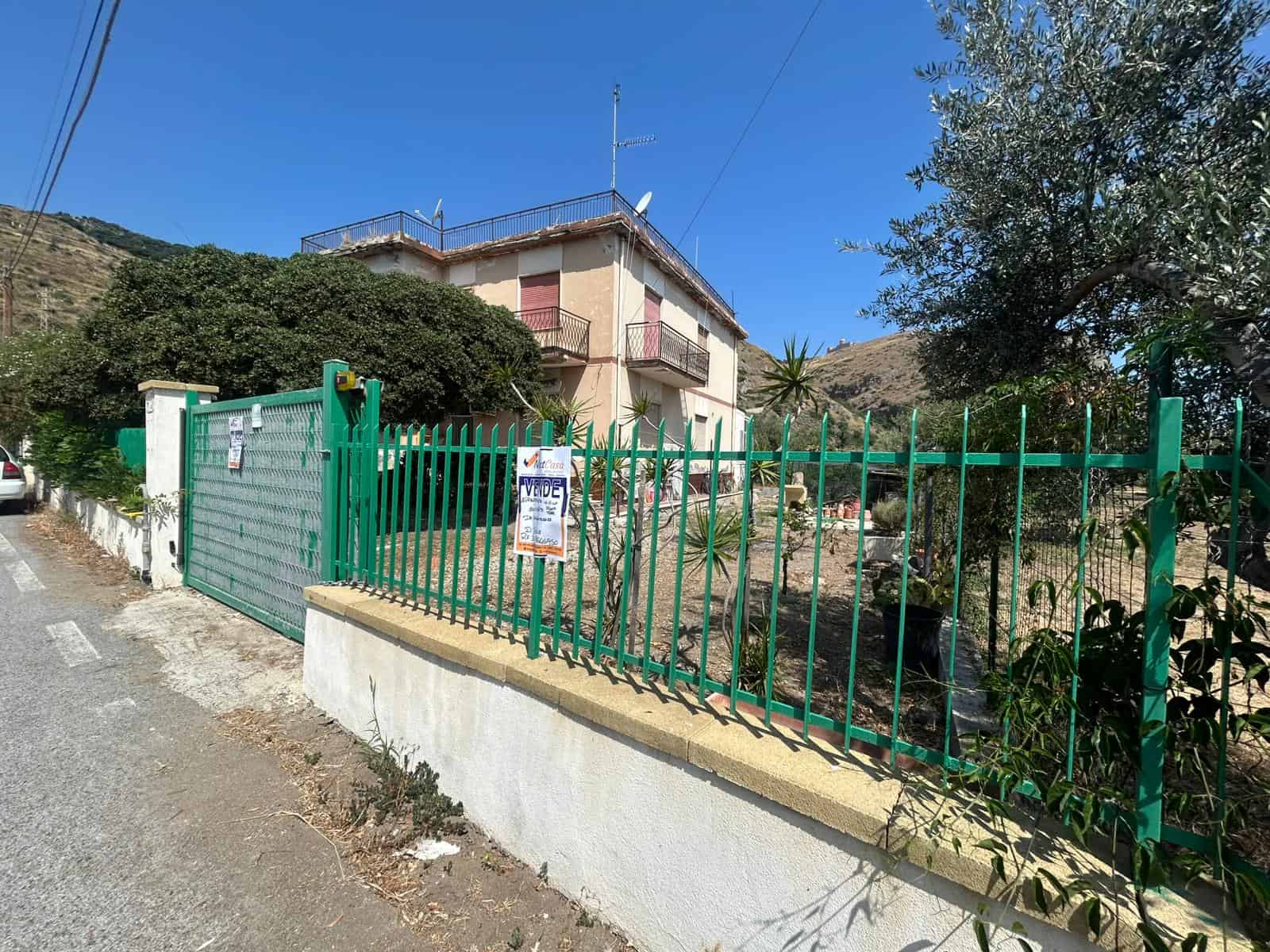 Una casa in vendita con giardino e cancello verde, situata in una zona collinare soleggiata, ideale per investimenti immobiliari e residence tranquillo, con vista panoramica sulle montagne.