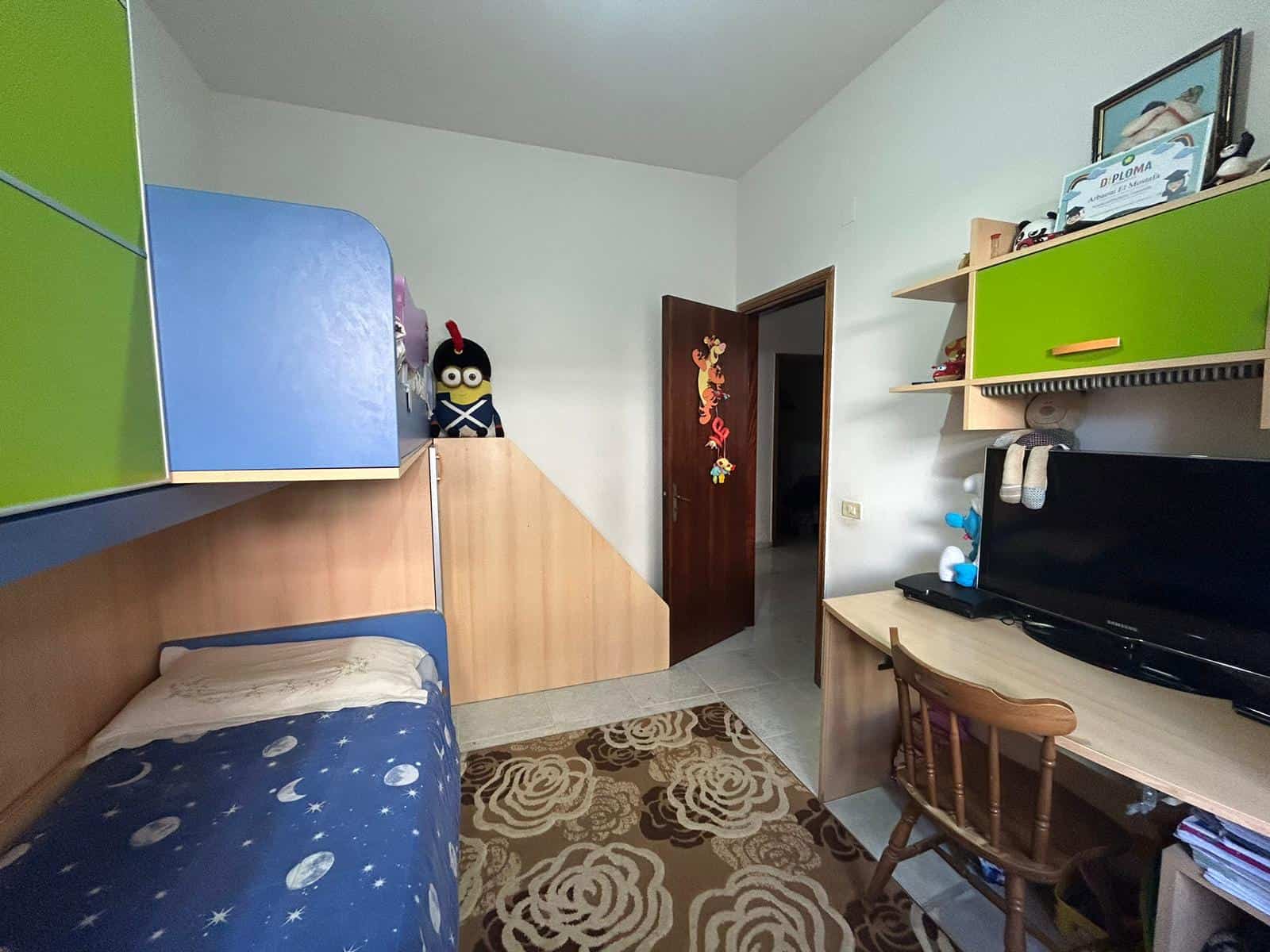 Letto singolo con copriletto con motivo spaziale, armadio verde e blu, scrivania con televisore e sedia in legno, decorazioni colorate sulla porta, stanza per bambini, arredi funzionali, atmosfera familiare.