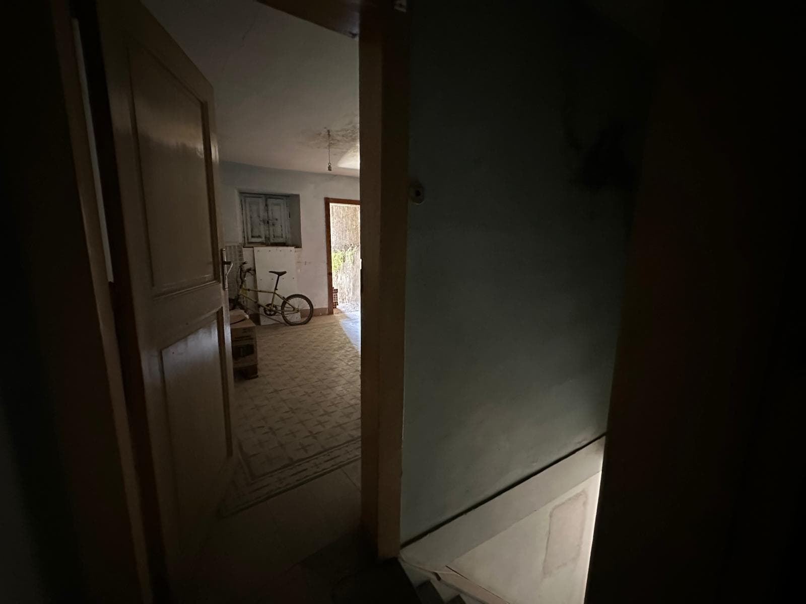 Vestibolo di casa in condizioni rustiche, con luci naturali che filtrano da una finestra e una porta in background, con biciclette e scatole come elementi di arredo temporaneo, ambiente semplice e funzionale.