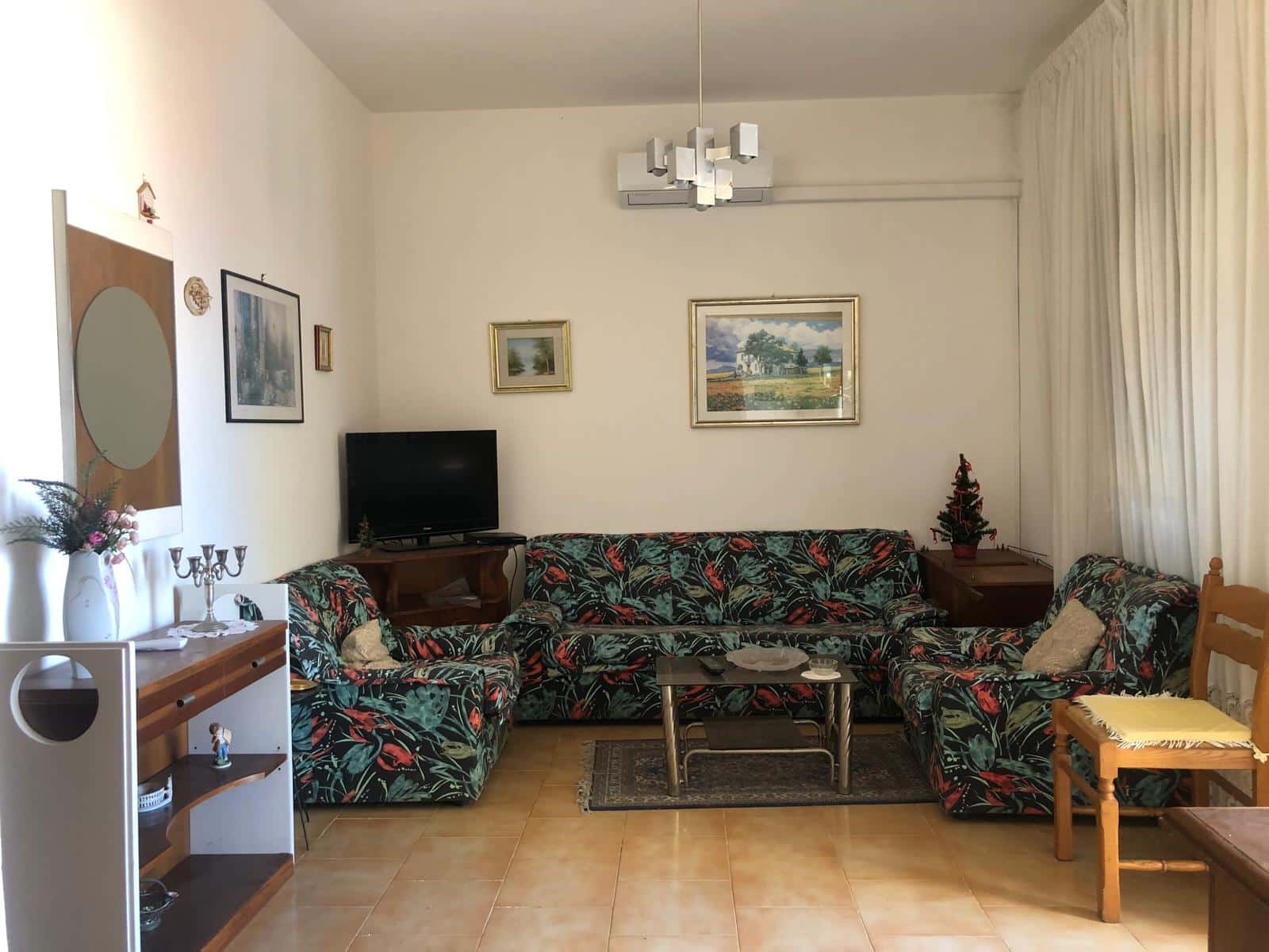 Ampia stanza soggiorno con divani a fiori, TV, quadri alle pareti e decorazioni domestiche, ideale per relax e socializzazione in ambienti accoglienti e luminosi.