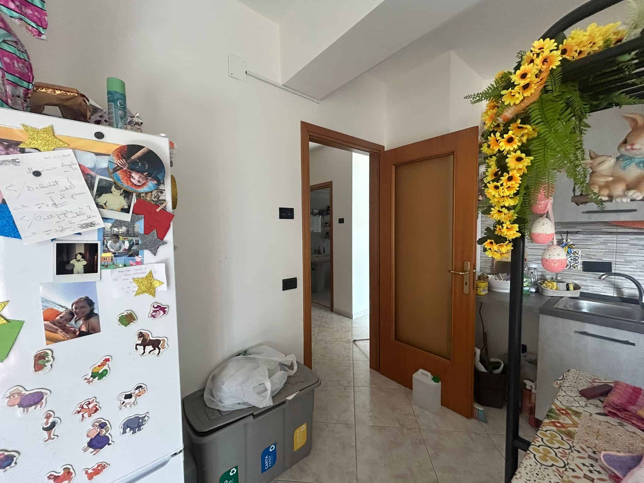 Altre testo descrittivo dell'immagine in italiano (max 473 caratteri): Interno di una cucina domestica con frigorifero decorato con foto e magneti, utensili e prodotti sugli scaffali, porta in legno che conduce ad altre stanze, decorazioni floreali e decorazioni di gatti, piano di lavoro con oggetti vari, ambiente vivace e accogliente.