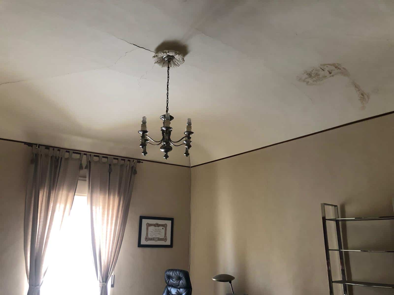 Vecchio soffitto con evidenti crepe e macchie, dettagliato con lampadario d'epoca e tende beige in una stanza abitativa. Interventi di ristrutturazione necessari per migliorare l’estetica e la sicurezza degli ambienti interni.